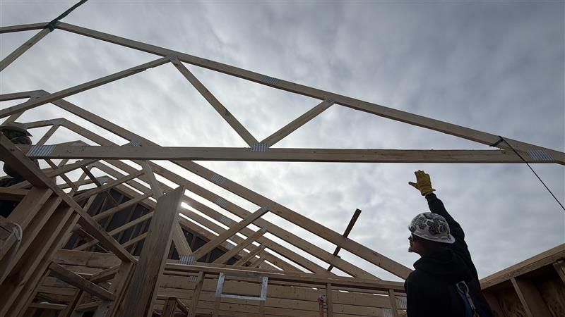 Construction worker guiding a wooden roof truss into place on a partially built house, against a cloudy sky.