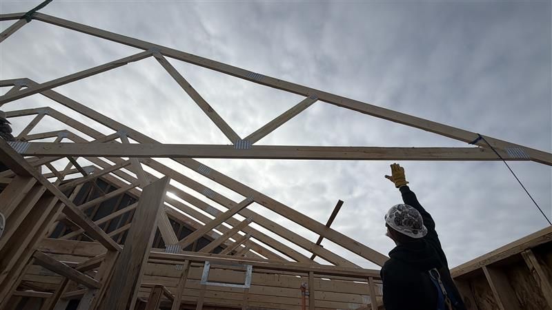 Construction worker, reaching, under wooden roof trusses against a cloudy sky.