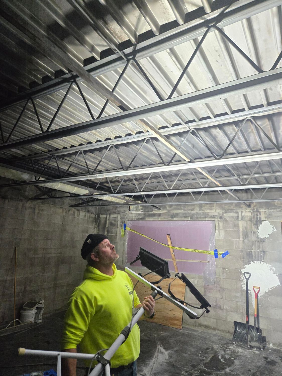Man in yellow shirt examines ceiling trusses in a construction site; concrete walls.