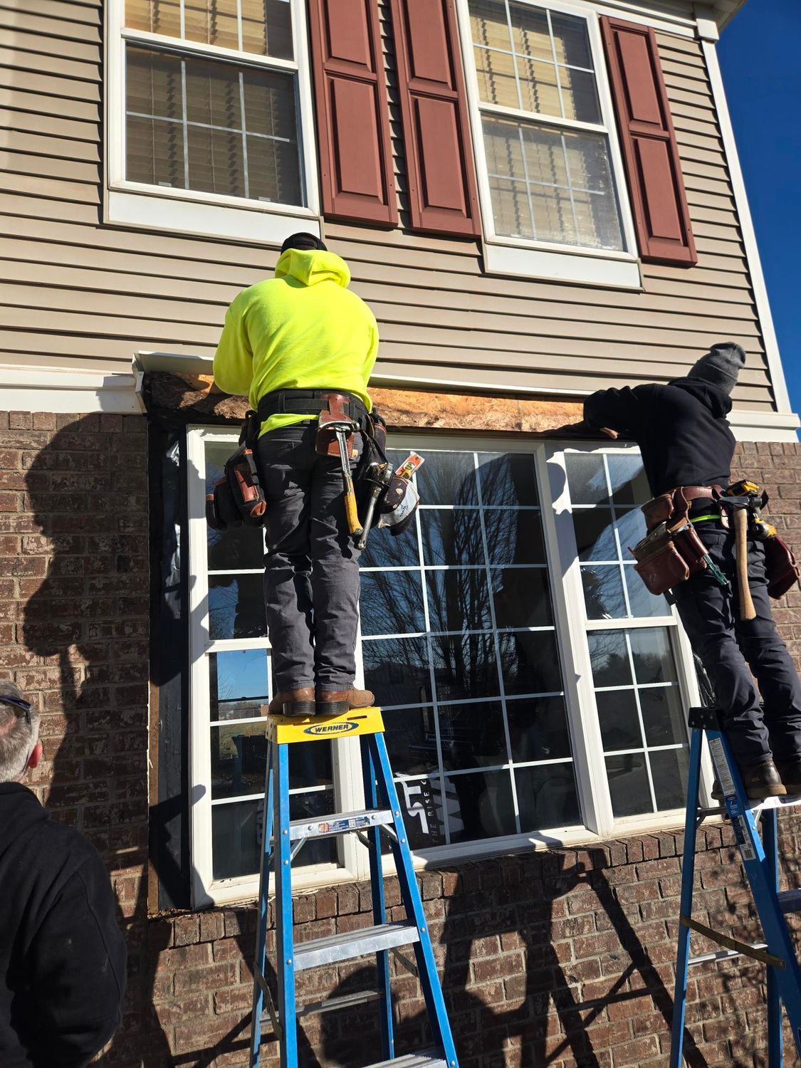Two construction workers installing a window, one on a ladder, working on the brown trim outside a brick home.
