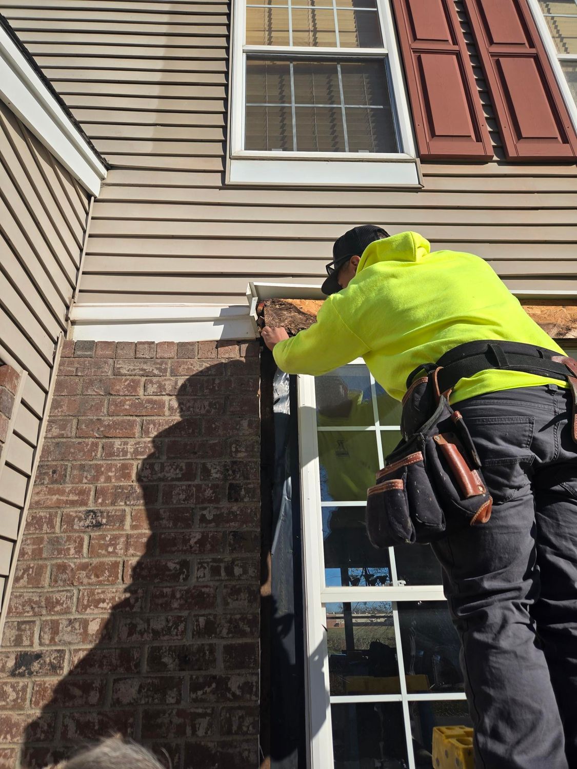 Construction worker in a yellow hoodie, on a ladder, working on a window frame. Brick and siding on the house.
