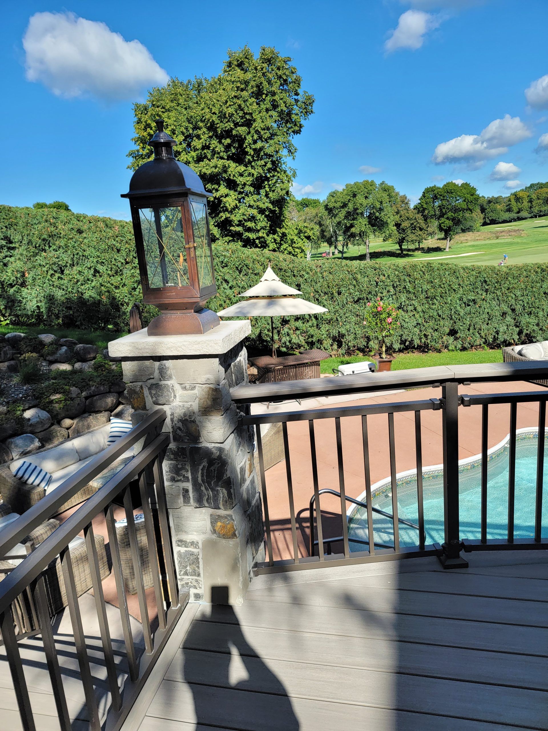 Outdoor deck with a stone pillar, a lamp, and a view of a pool and greenery under a blue sky.