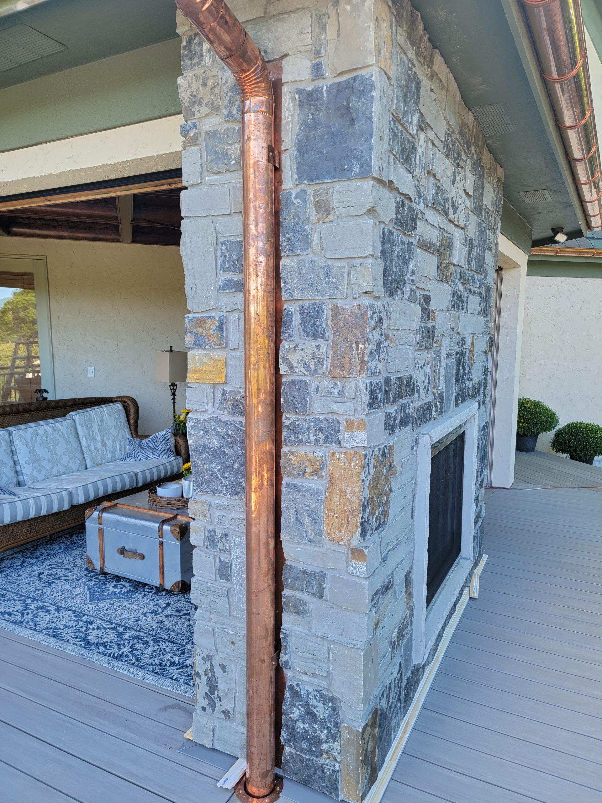 Copper rain gutter on stone wall of an outdoor fireplace, part of a patio with a couch.