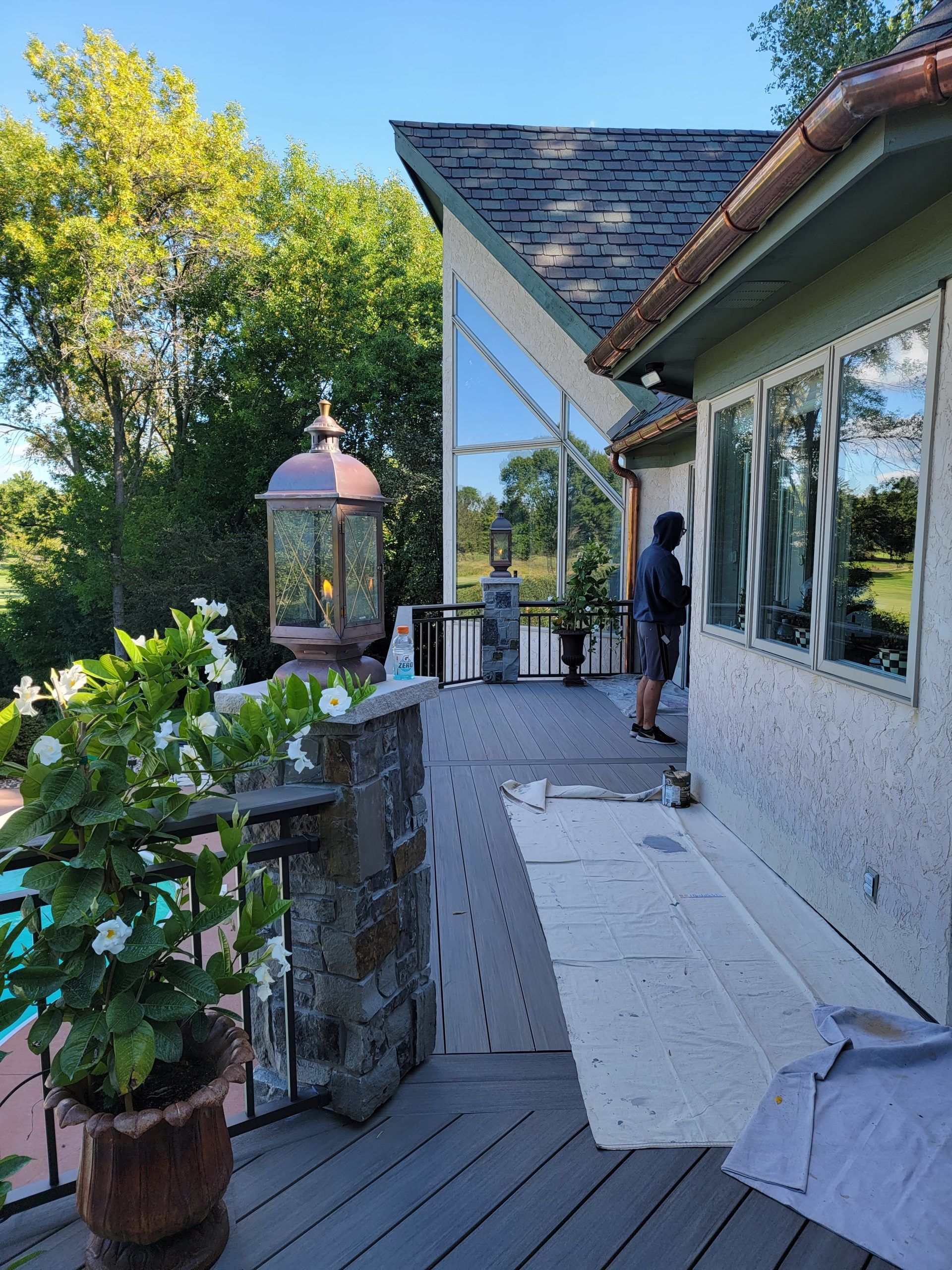 Deck with potted plants, pool, and house with large windows. Person near the building.