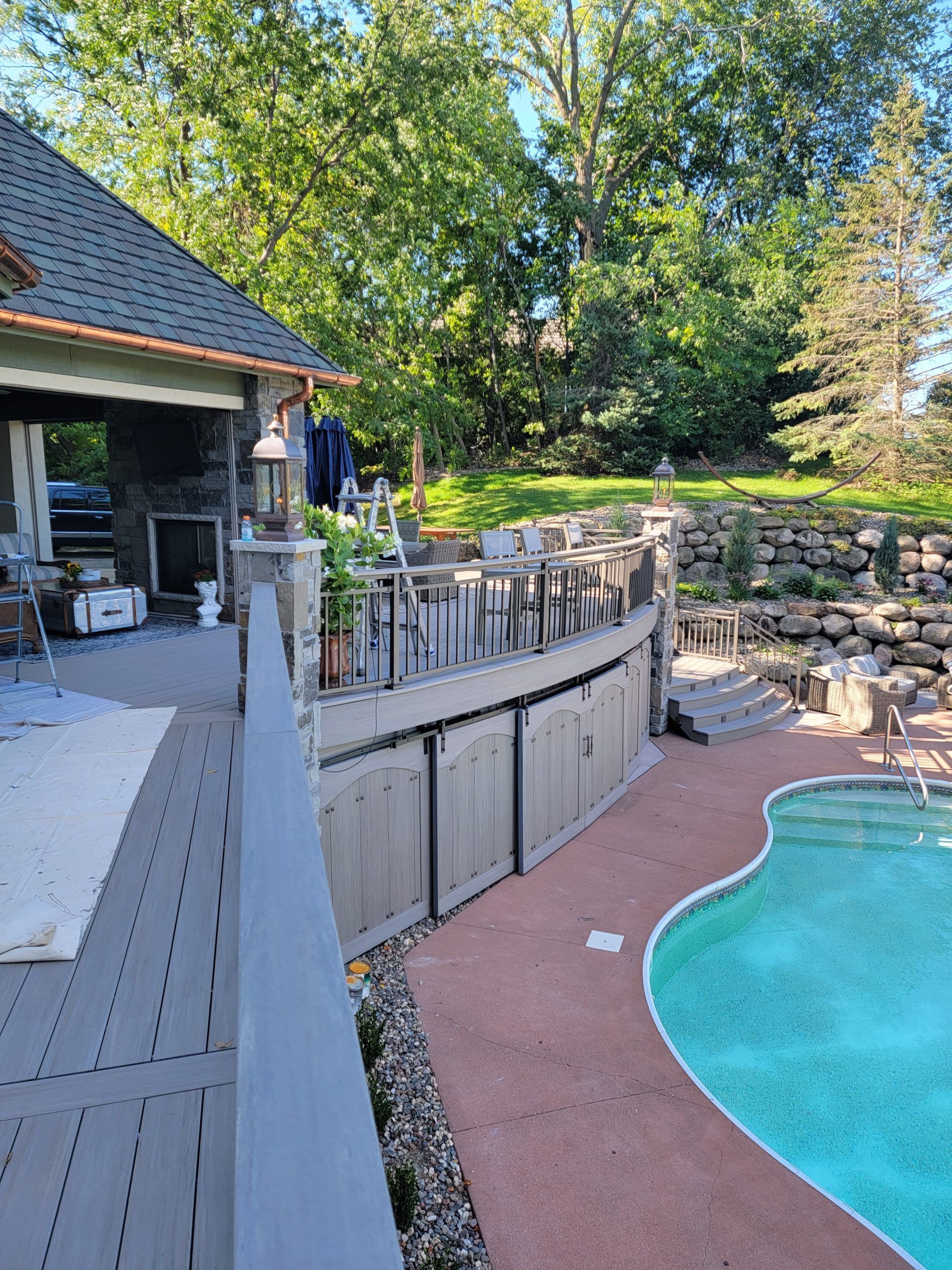 Backyard deck with pool and outdoor seating. Trees and house with stone chimney in the background.