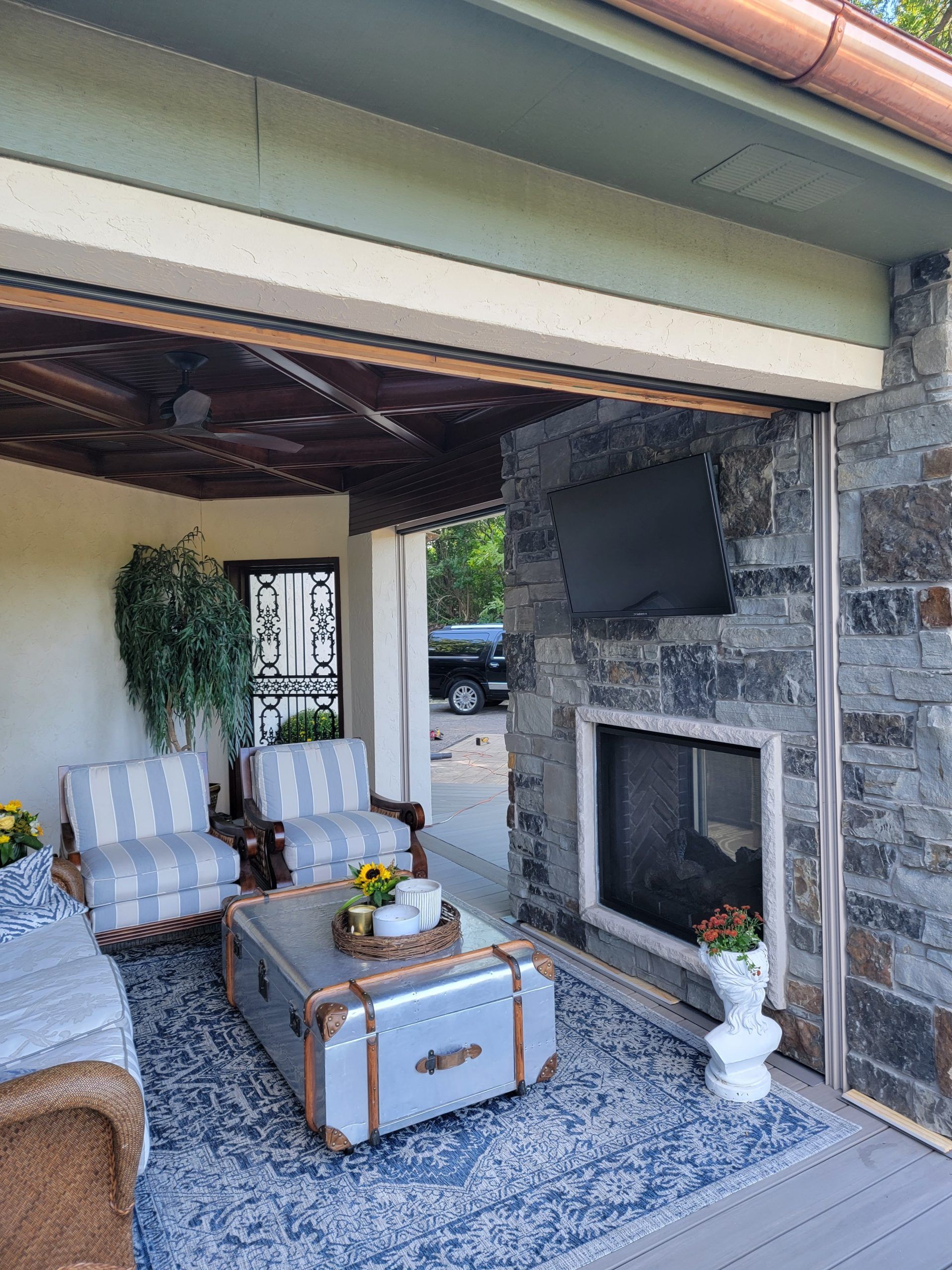 Outdoor living space with fireplace, TV, seating, and decorative trunk on blue rug.