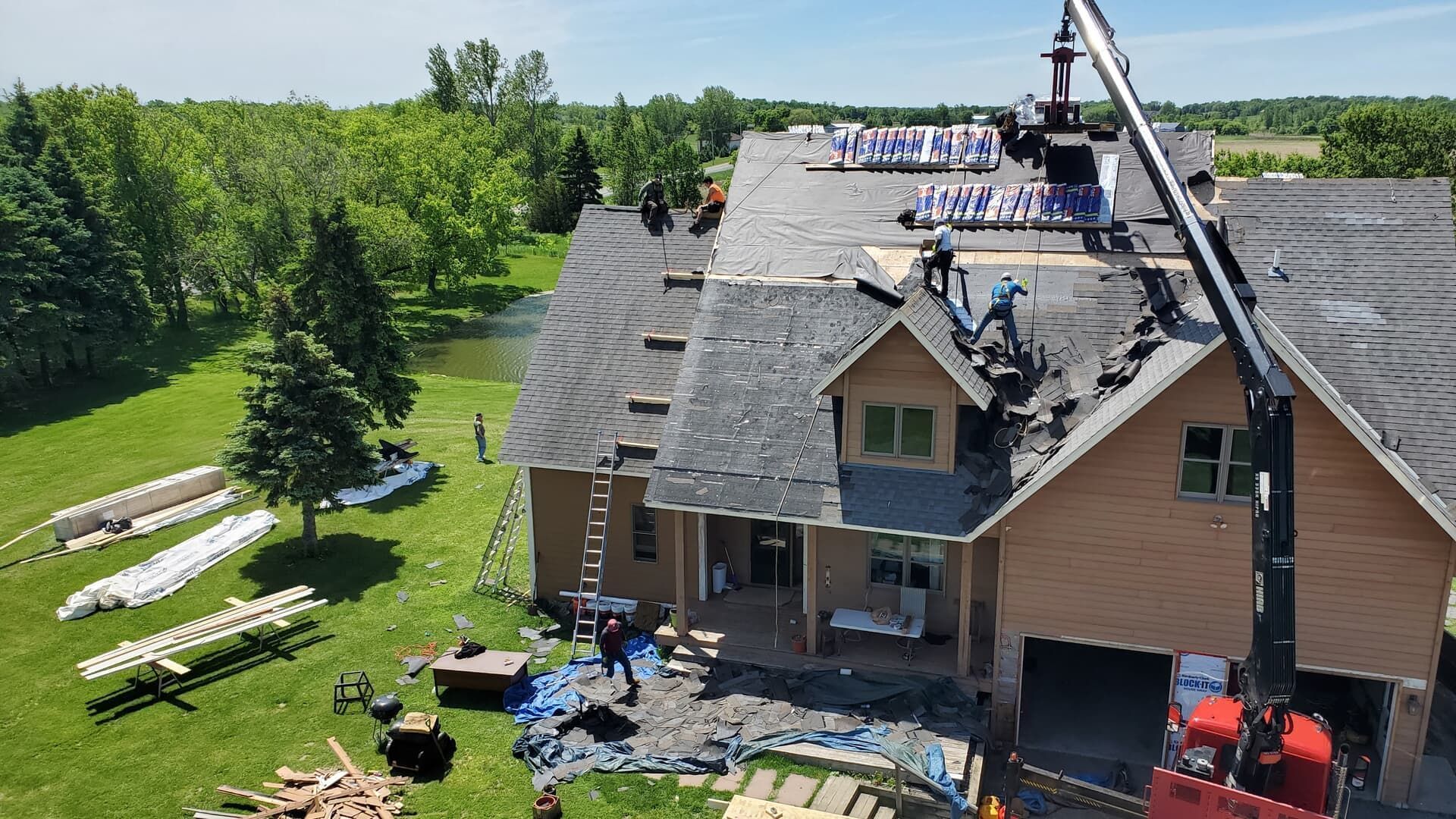 Roofers working on a house with a crane. Blue sky, green trees, and materials on the ground.