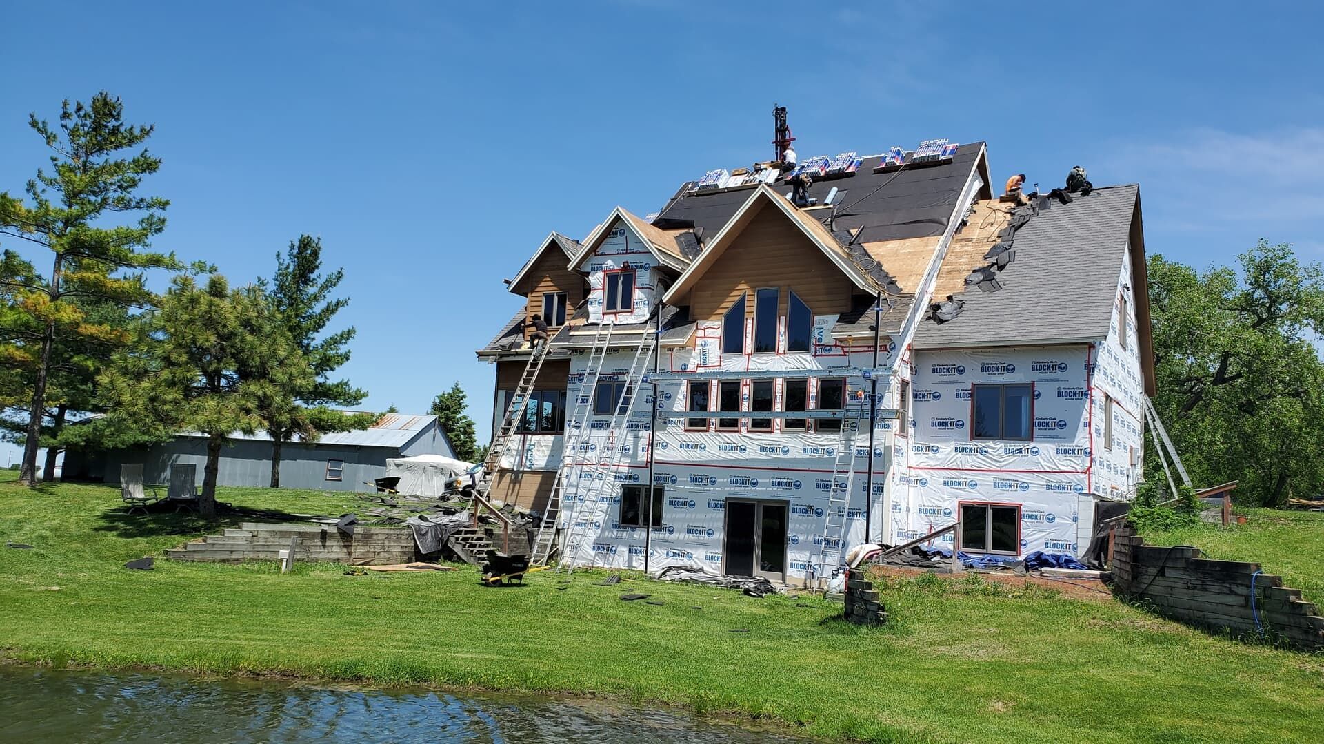 House under construction with missing roof shingles and protective wrap; surrounded by grass and trees.