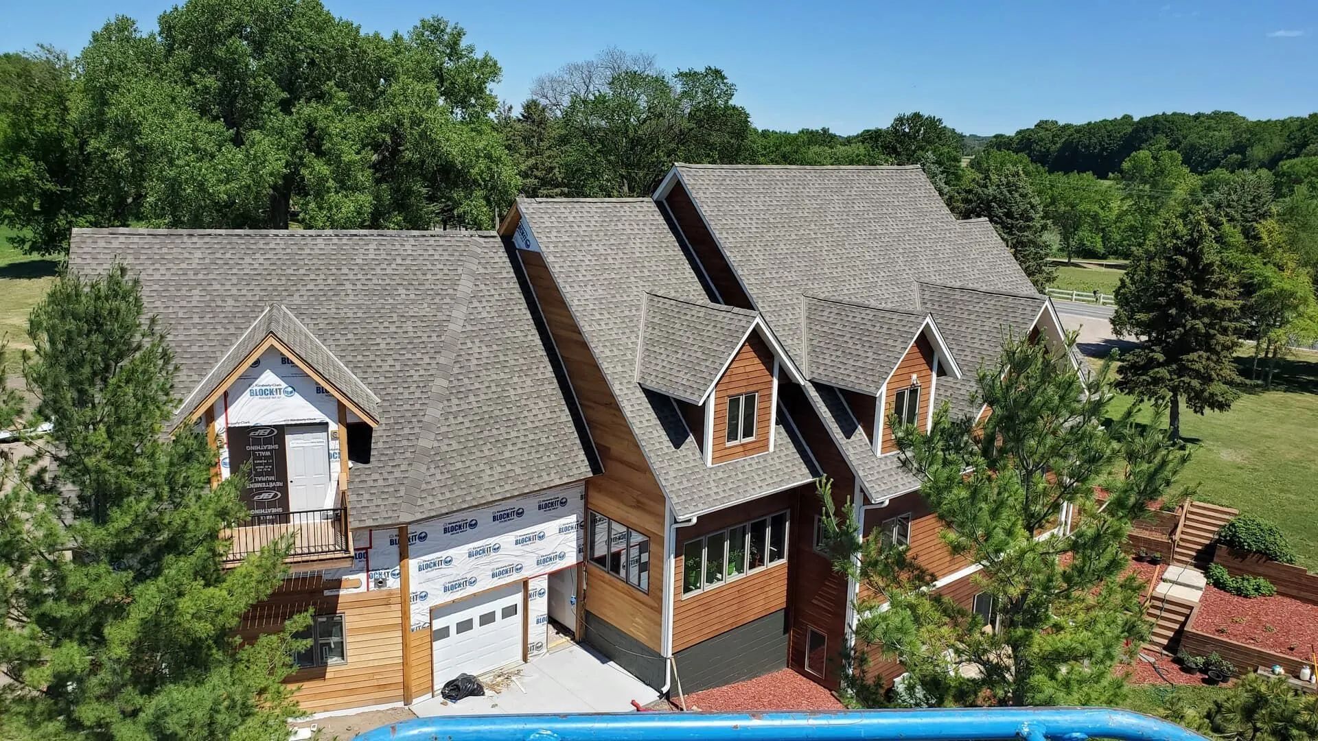 Aerial view of a large gray-roofed house with wood accents beside green trees and a driveway.