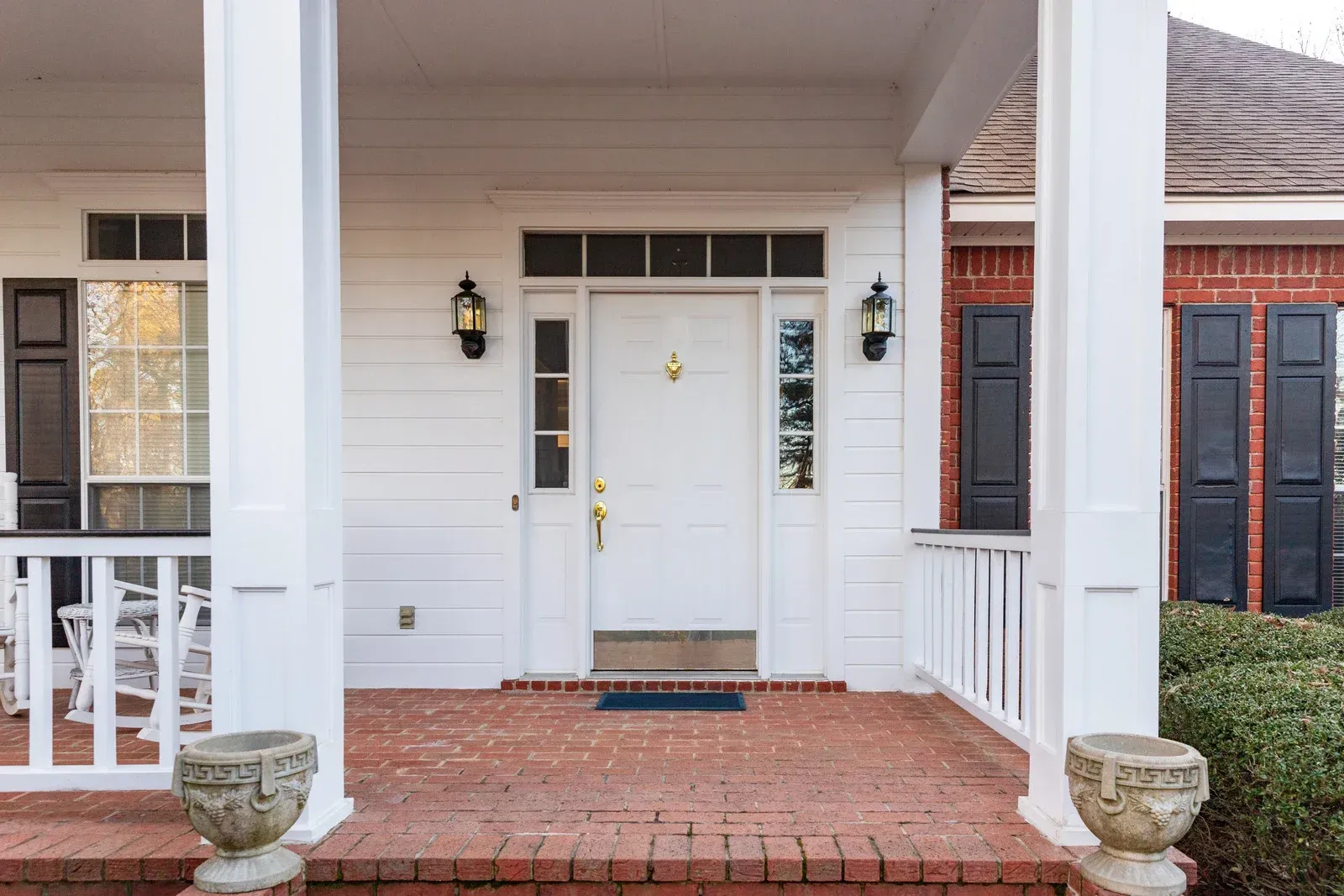 White house entrance with brick porch, white columns, door with sidelights, black shutters.