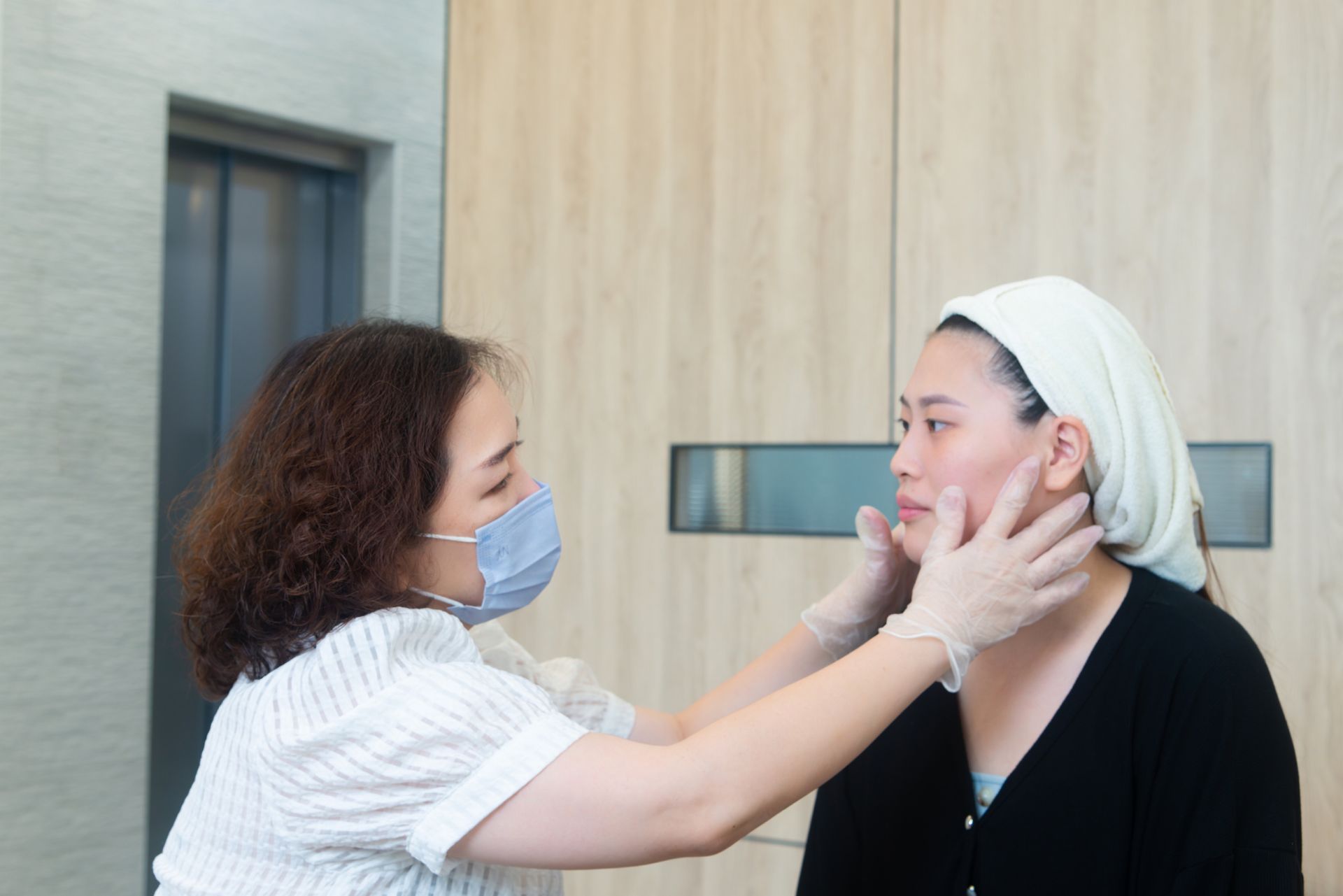 兩名戴著口罩的女子在室內面對面站著，其中一人輕輕幫另一人調整口罩。