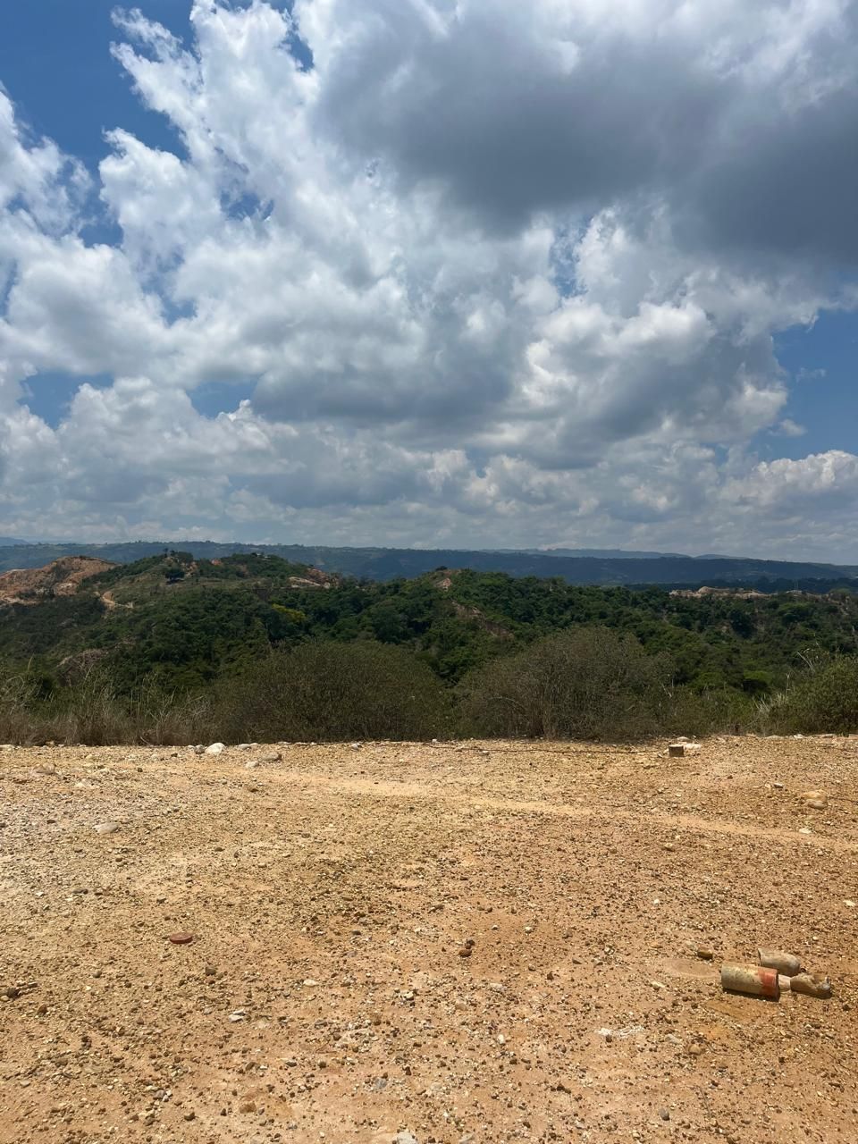 Un campo de tierra con árboles al fondo y un cielo azul con nubes.
