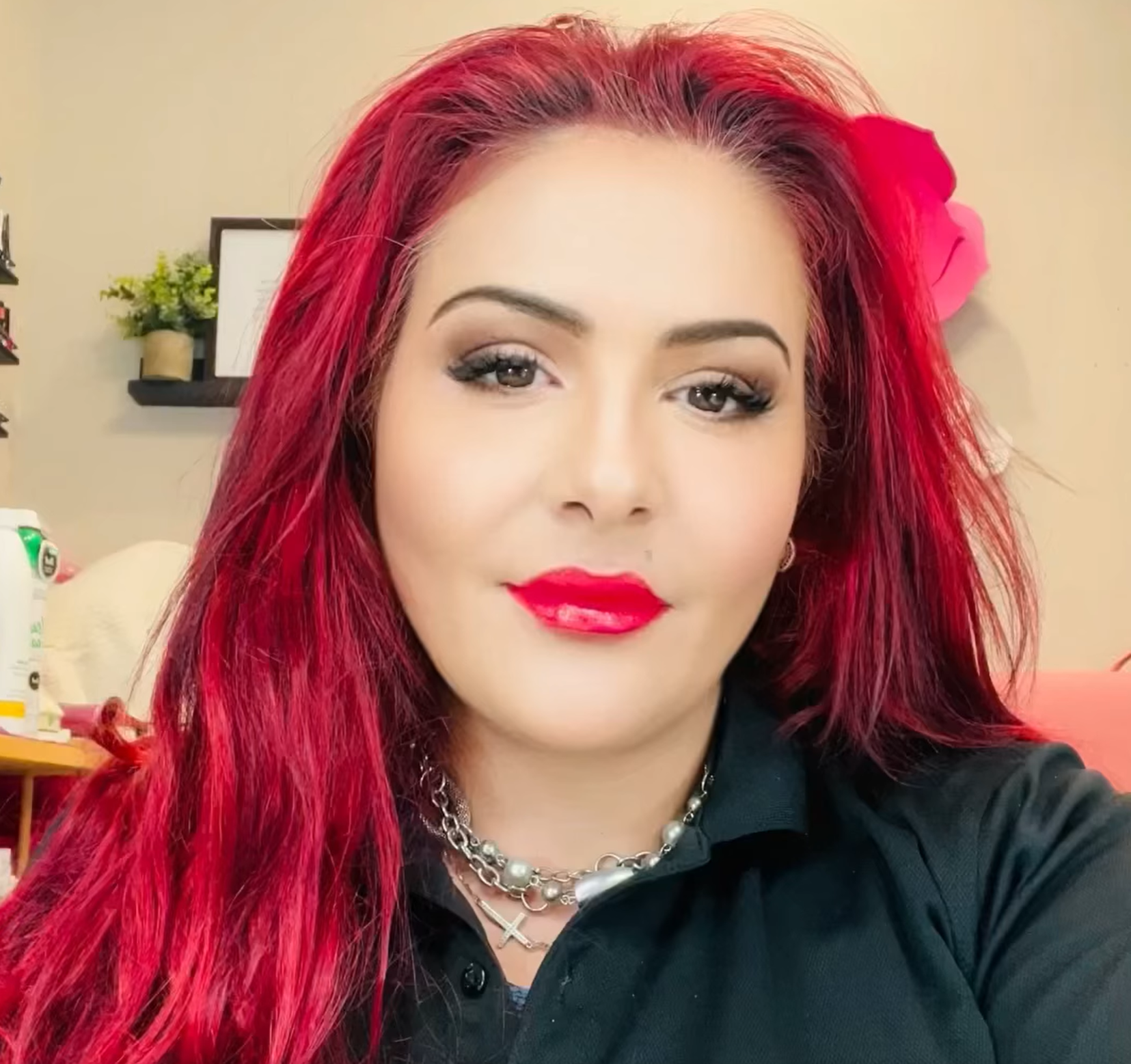 Woman with red hair, red lipstick, silver necklace, wearing a black shirt, soft glam makeup in a well-lit room.