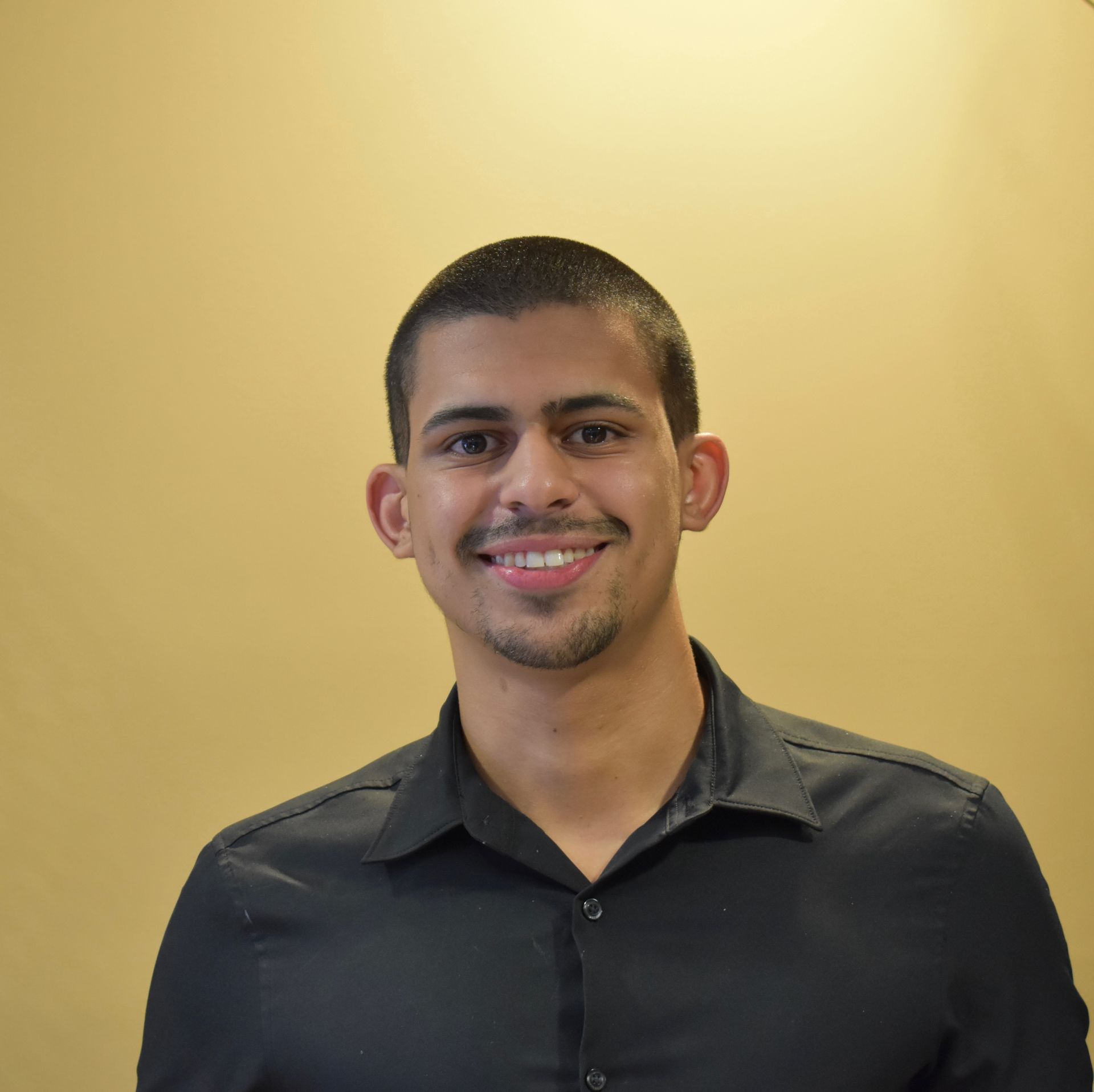 Young man with short dark hair, smiles, and wears a black button-down shirt in front of a yellow wall.