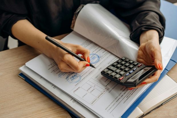 Person with red nails using a pen to write on paperwork, a calculator is nearby.