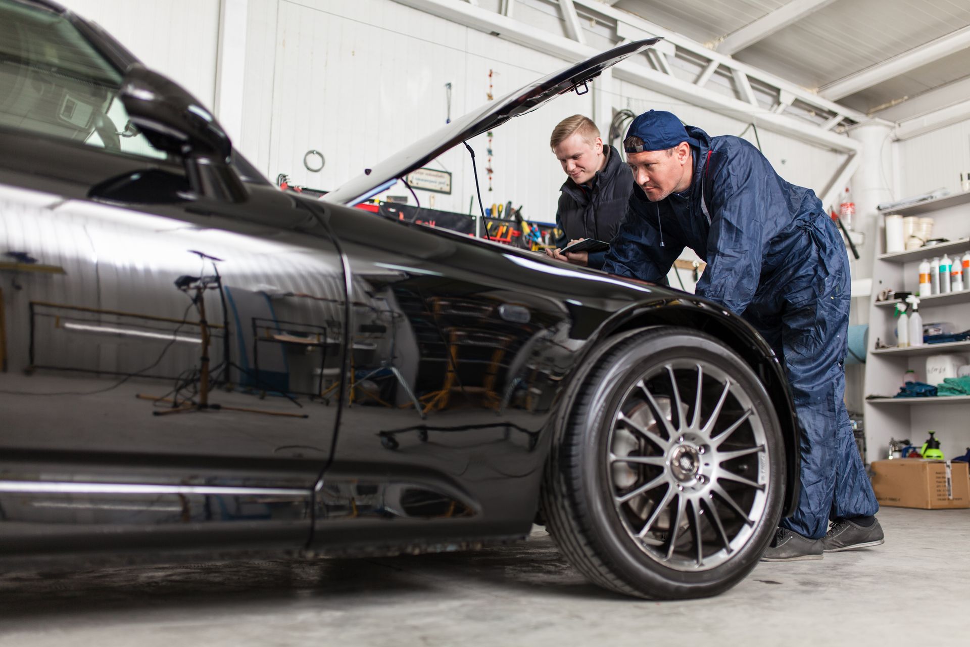 Two mechanics examining a black car with the hood open in a garage.