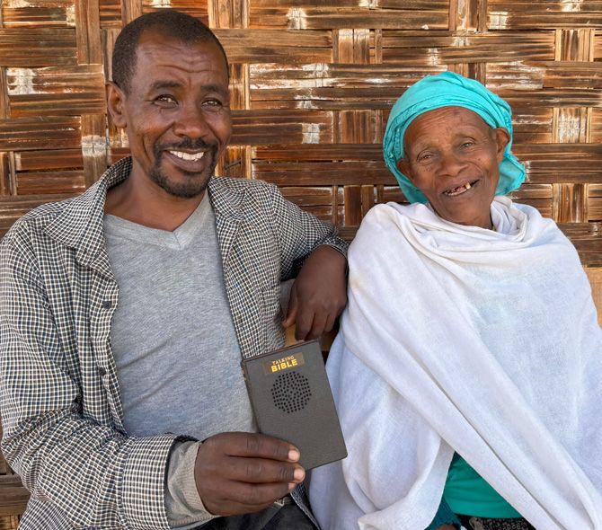 Man and woman smiling, holding a Talking Bible, seated outdoors. Man in jacket, woman wrapped in white.