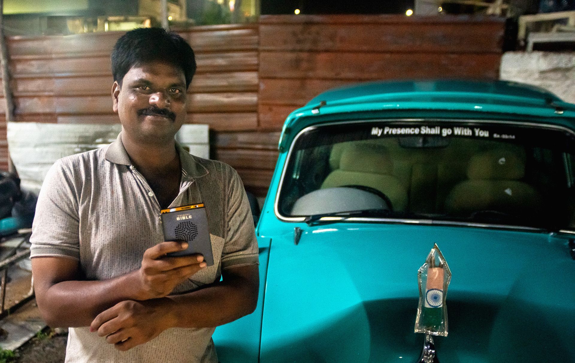 Man holding a phone smiles next to a teal vintage car; night setting.