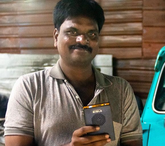Man holding a small electronic device, smiling. He wears a grey shirt and is in front of a teal vehicle.