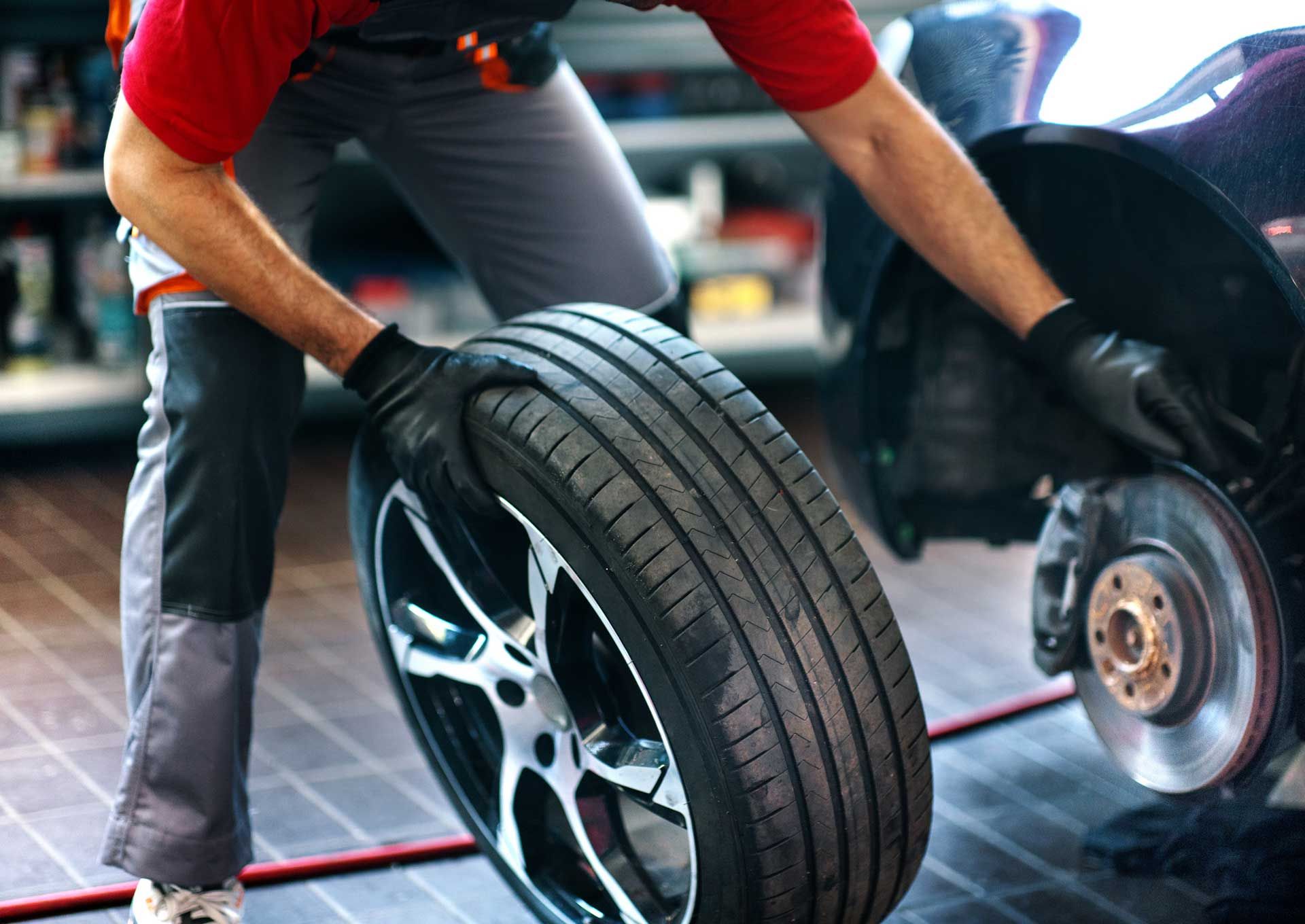 Mechanic in gray jumpsuit and red shirt changing a car tire; a tire is being lifted.