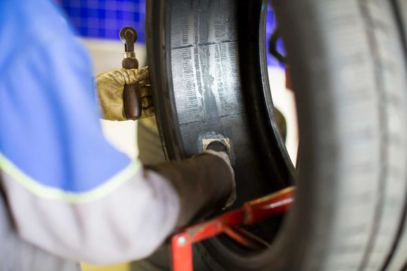 Person patching a tire in a garage, using a tool. The tire is black; the person wears gloves and a blue shirt.