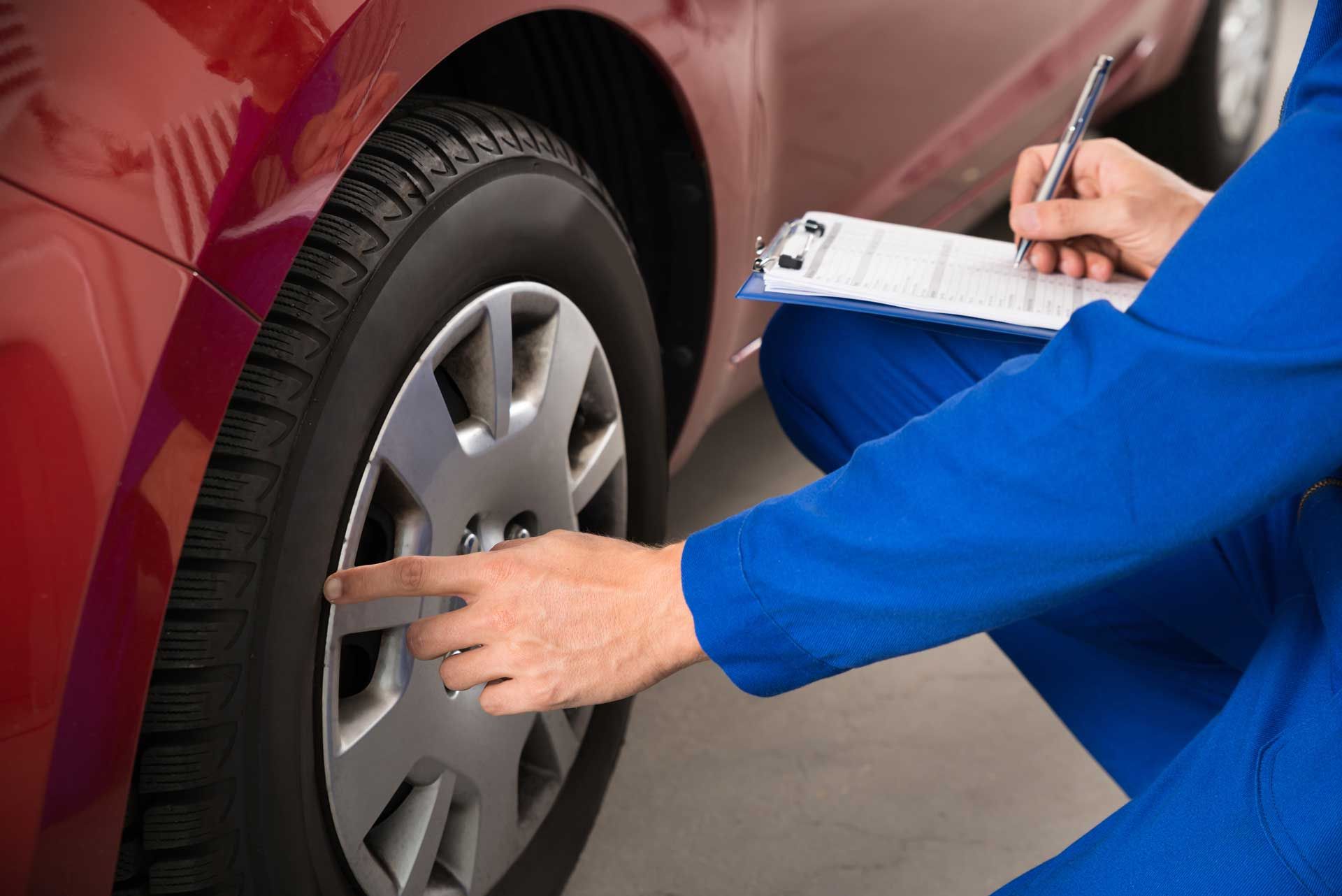 Mechanic in blue jumpsuit inspecting car tire, holding clipboard and pen. Red car in garage.