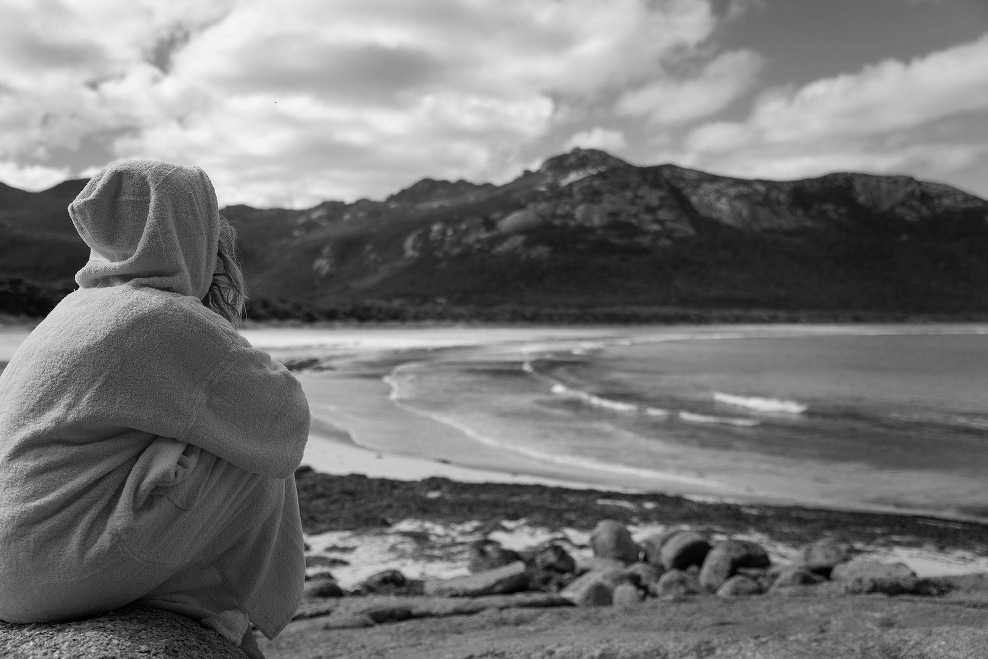 A black and white photo of a person sitting on a rock looking at the ocean, wrapped up in hoodie towel.