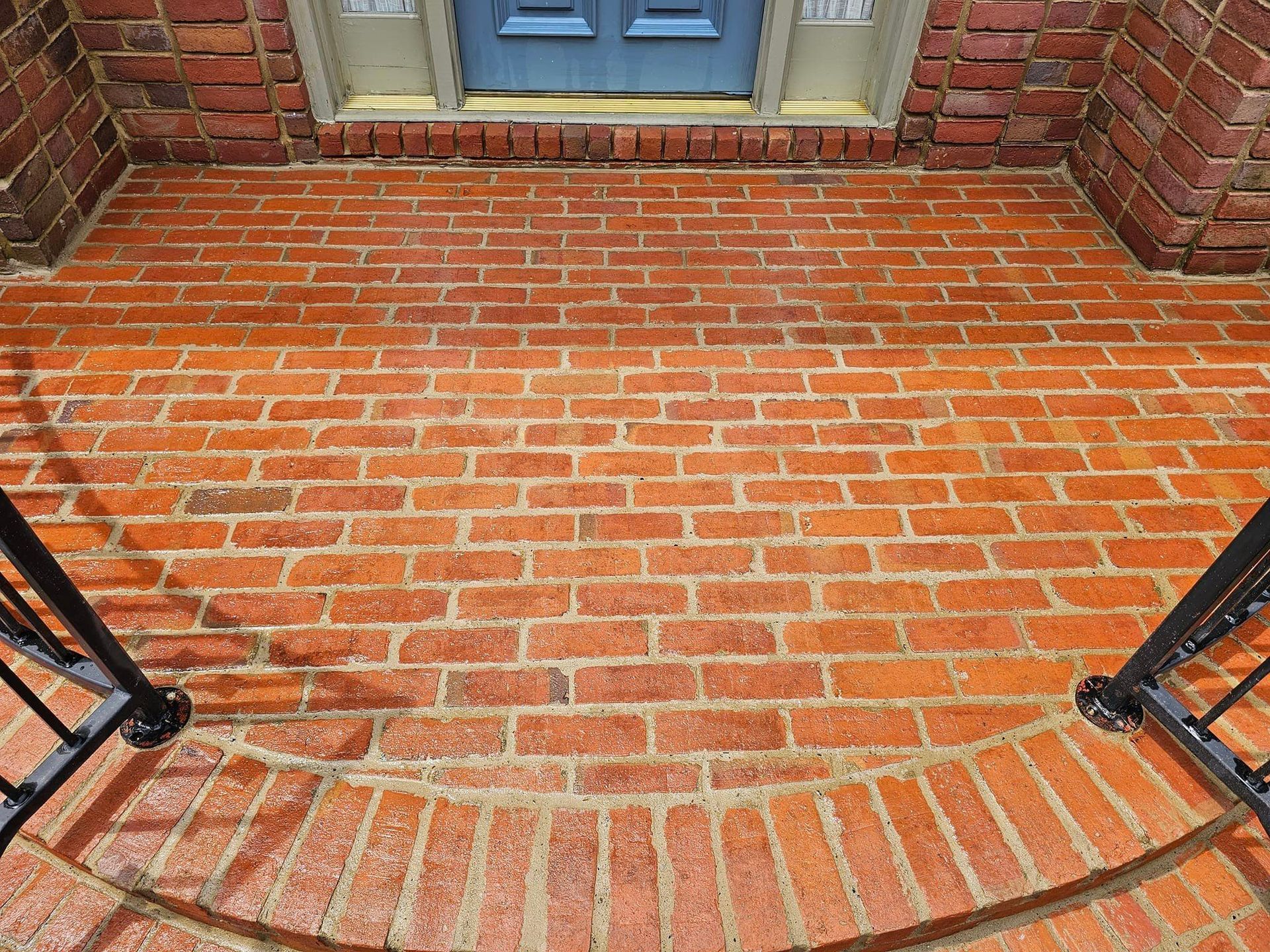 Red brick porch and steps leading to a blue door. Black railing on sides.