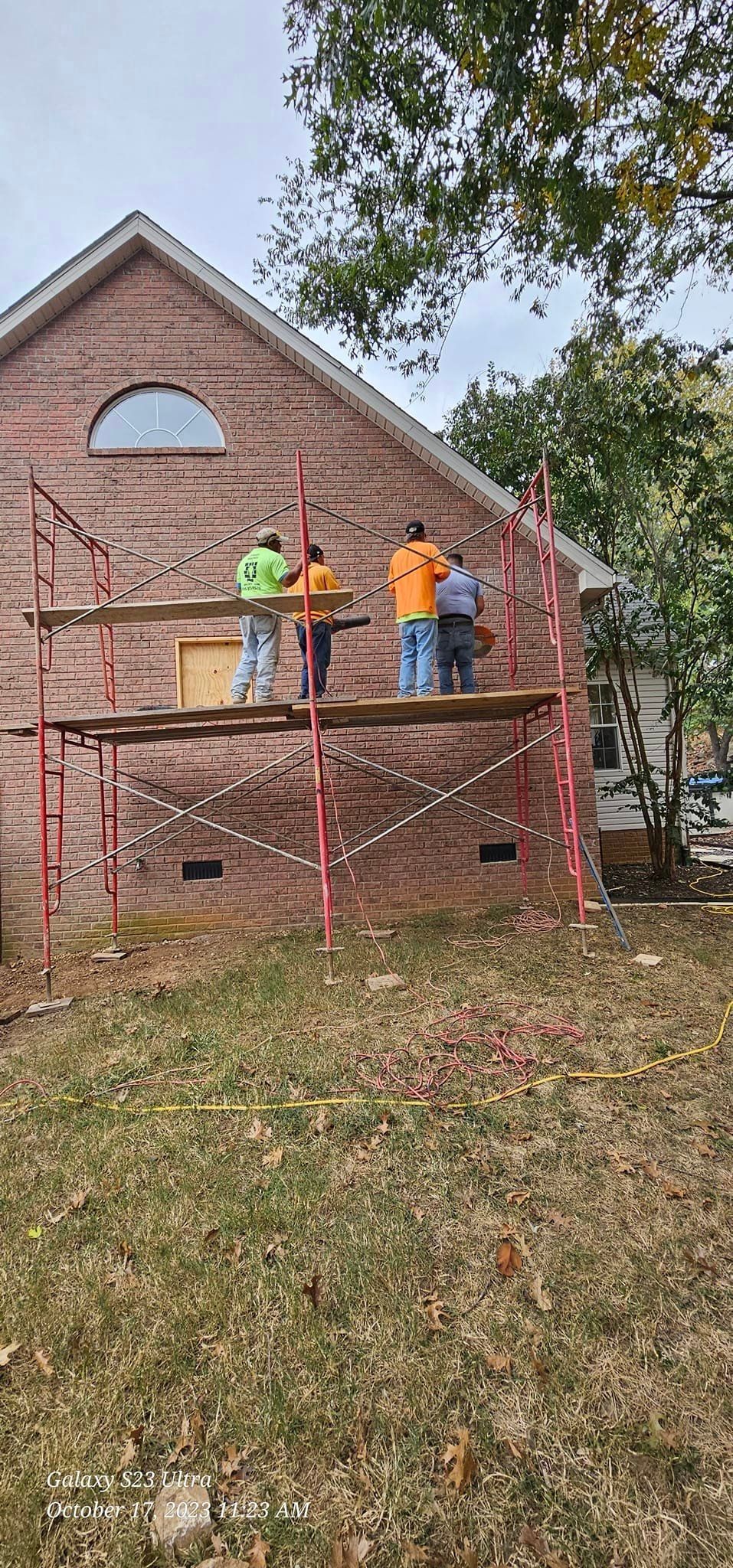 Construction workers on scaffolding against a brick building. Red scaffolding, green and orange shirts, cloudy sky.
