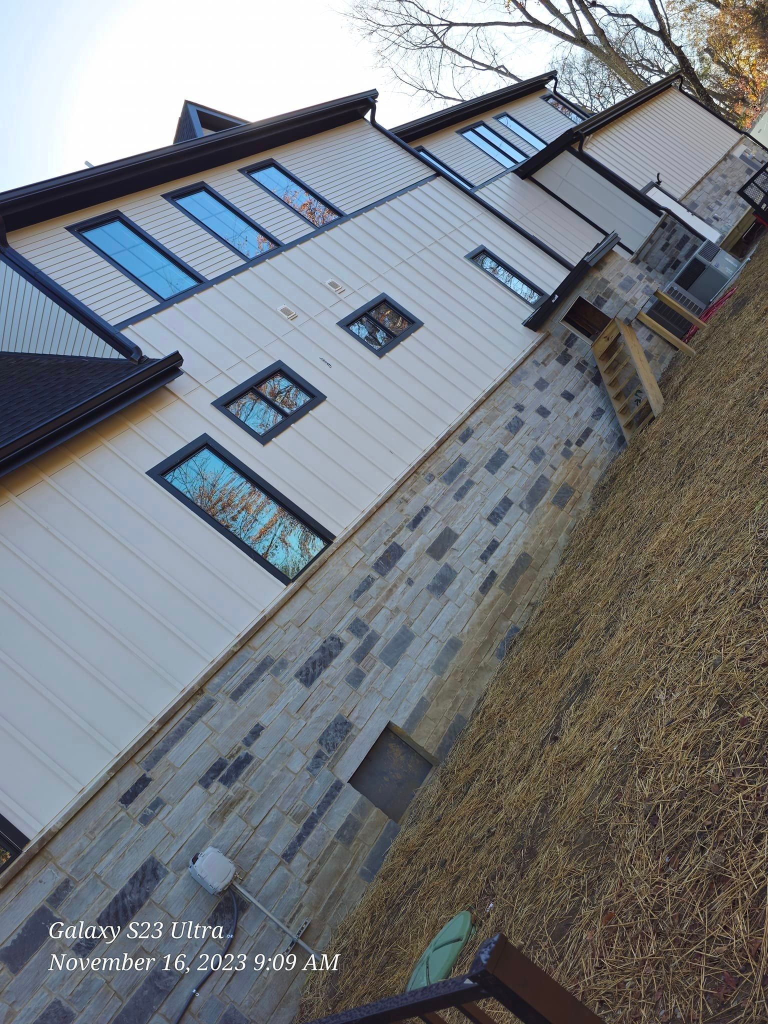 Two-story house with light siding, dark trim, and stone foundation. Windows reflect the sky.