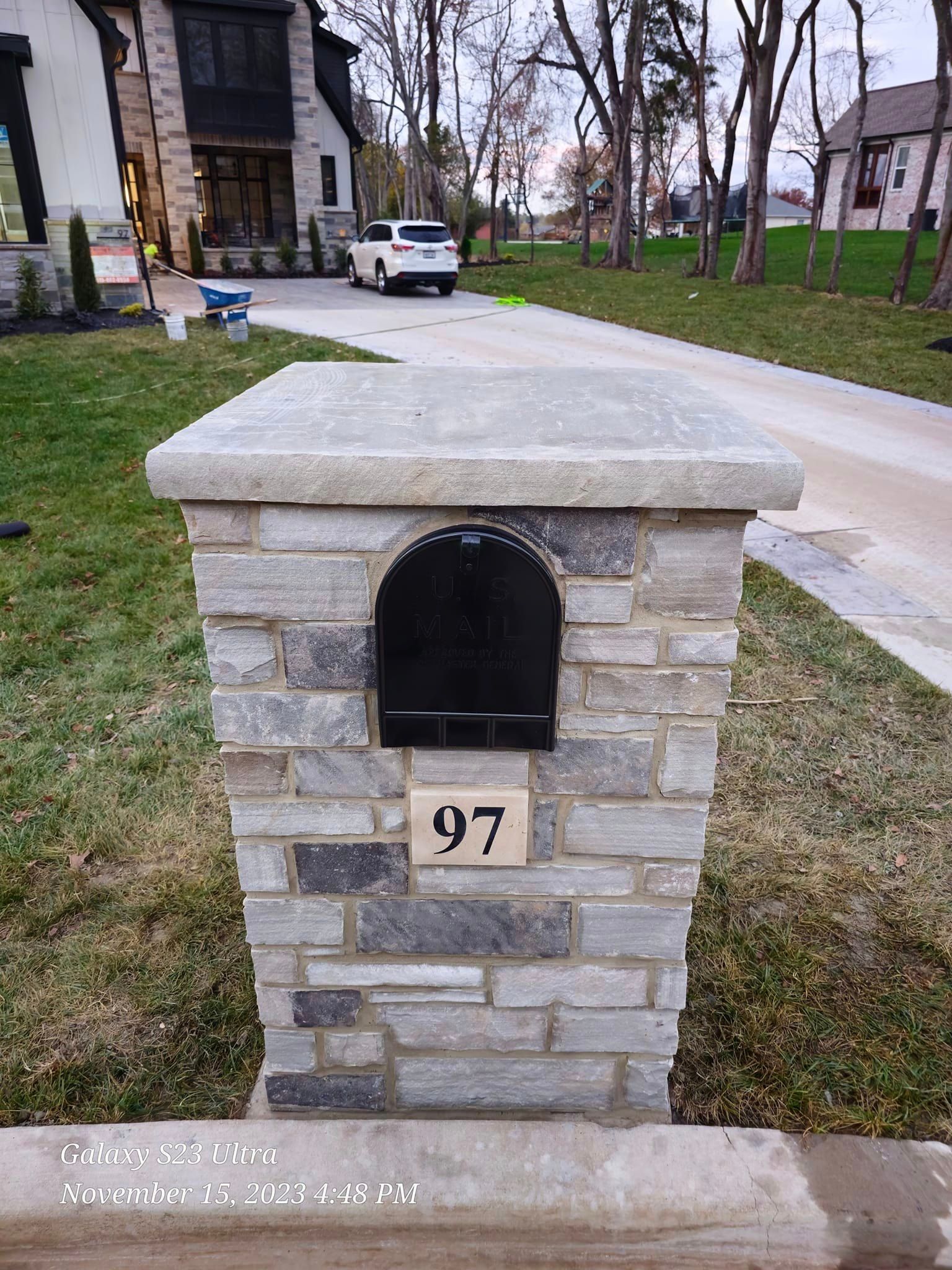 Stone mailbox with black door, number 97, sits on a lawn in front of a house and driveway.