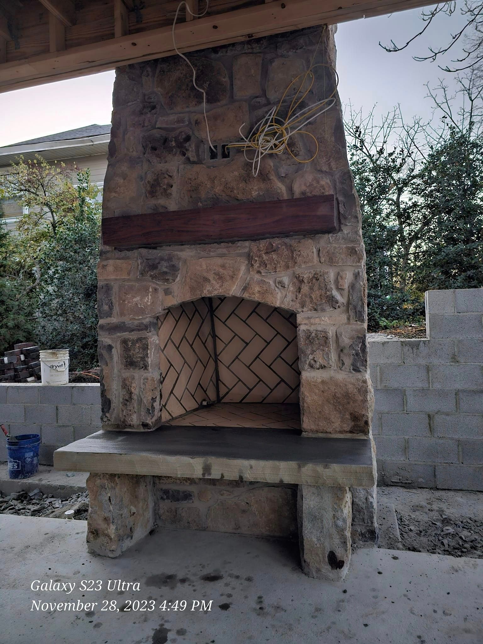 Stone outdoor fireplace with herringbone brick interior, wooden beam, and stone hearth.