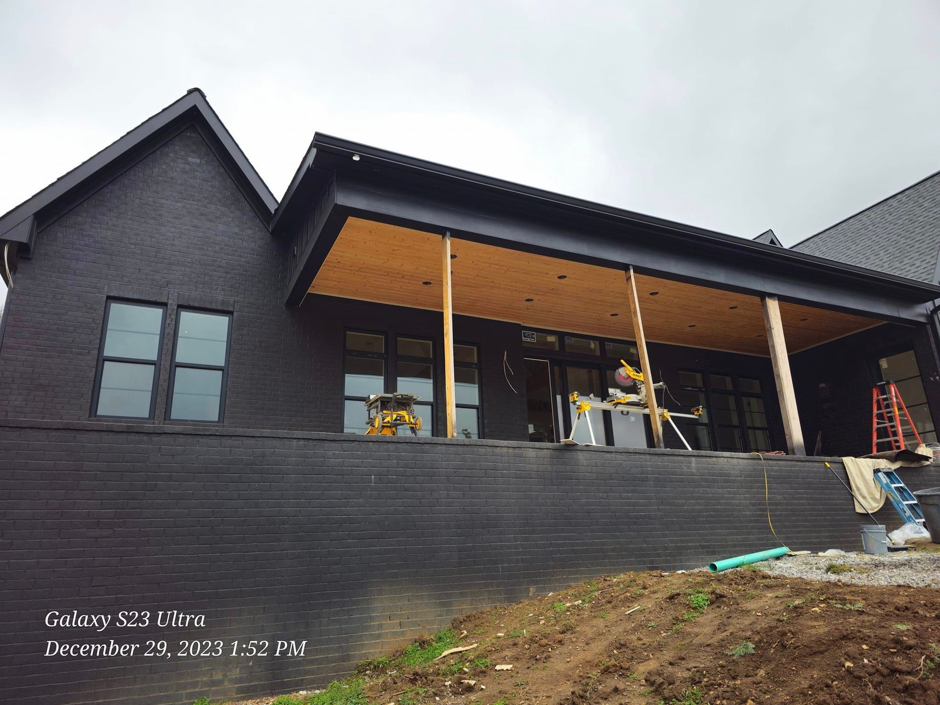 Black brick house under construction, wooden porch supports, cloudy day.