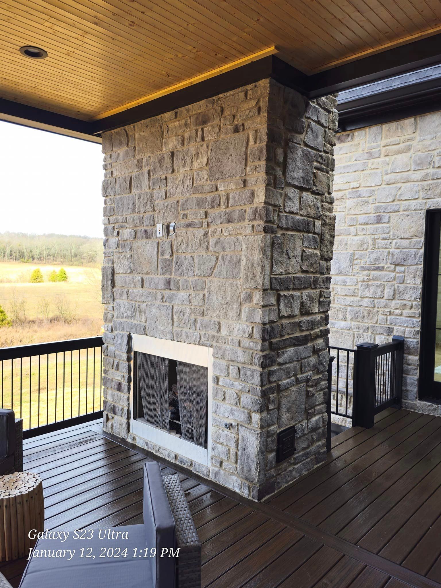 Outdoor fireplace built of gray stone on a wooden deck with a view of a field.