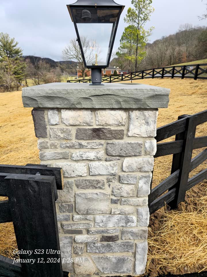 Stone pillar with lamp, gate and black fence in field with trees and hills.