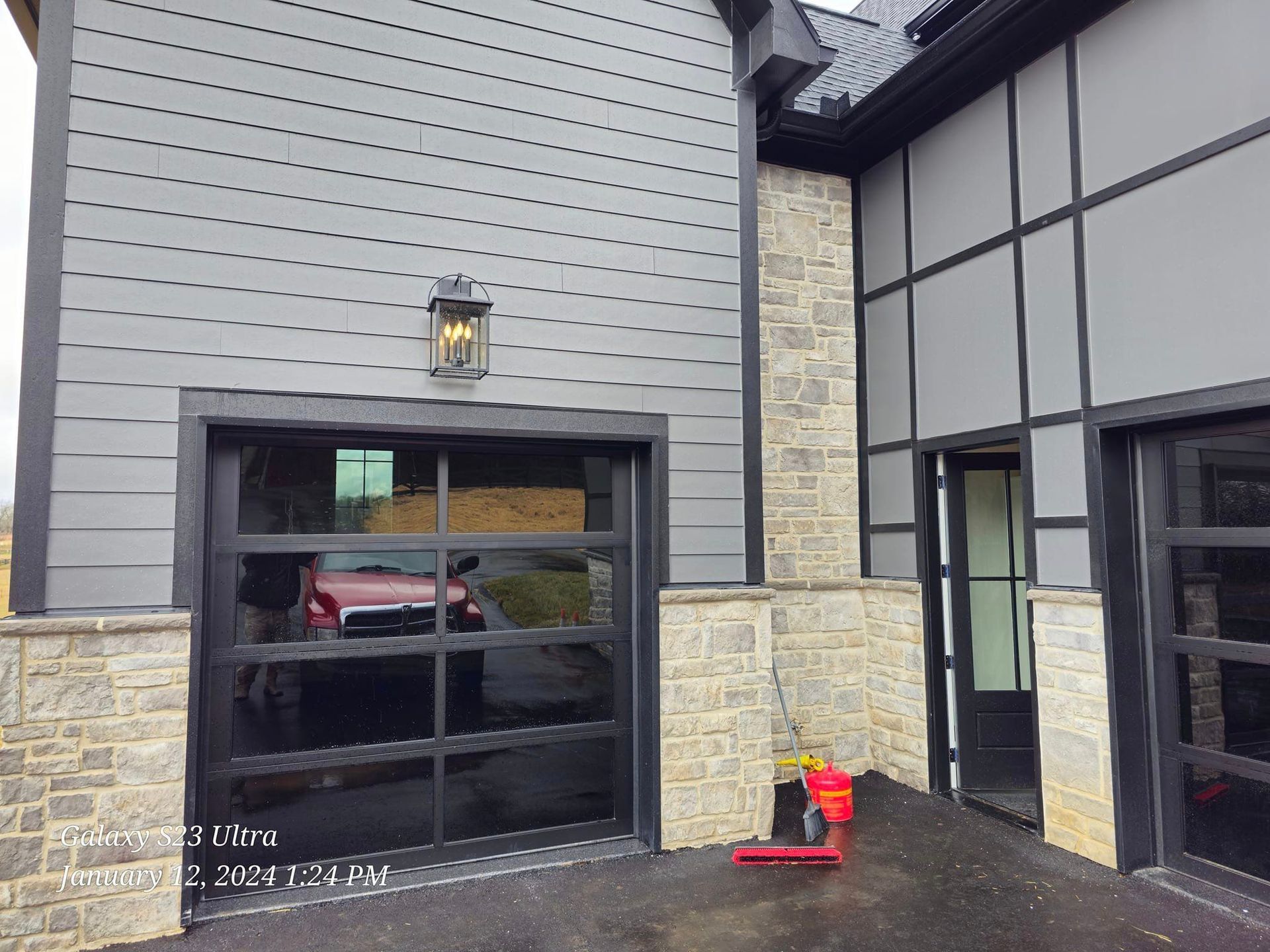 Garage with black-framed glass door reflecting a car, gray siding, stone accents, and an outdoor wall light.