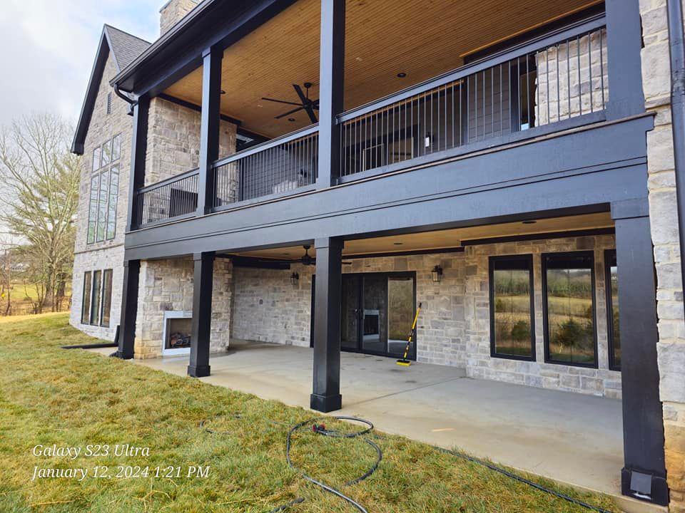 Exterior view of a two-story house with black trim and a stone facade, featuring a covered patio and balcony.