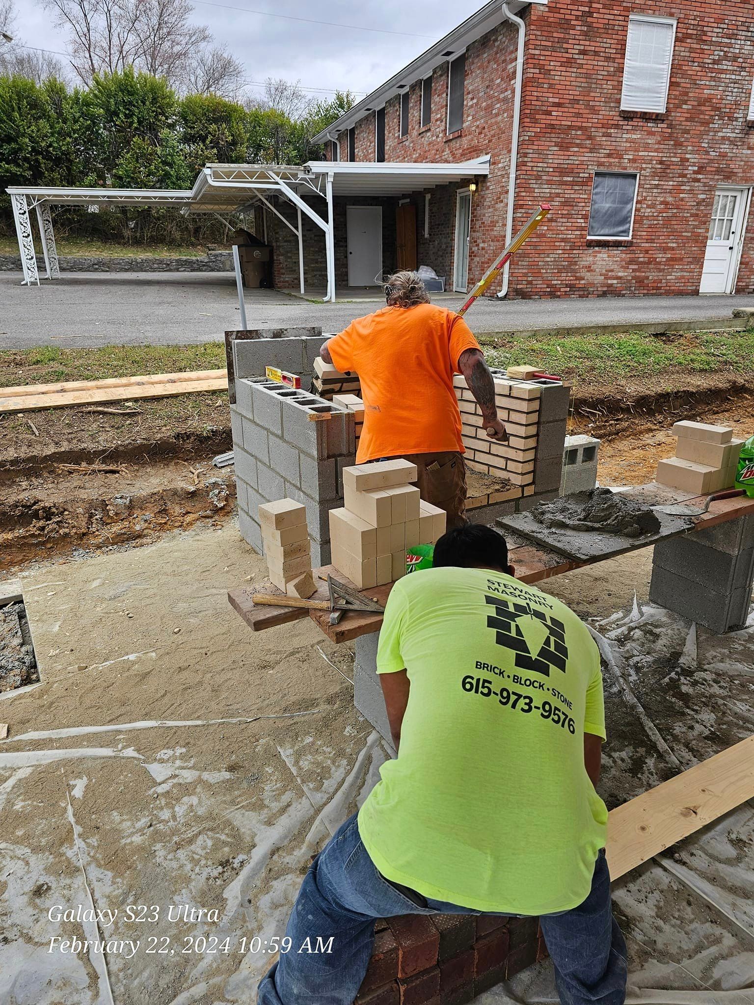 Construction workers building a wall with blocks, set in an excavation, near a brick building.