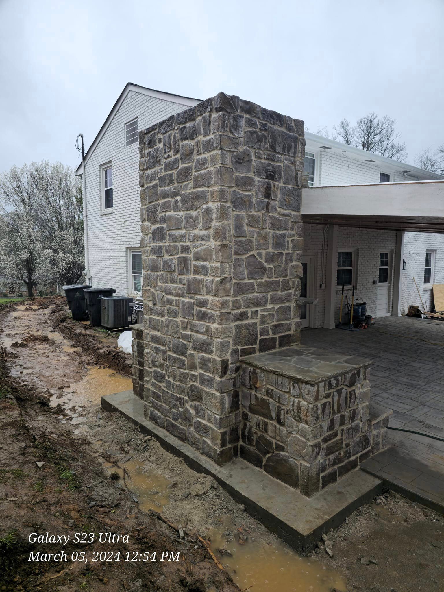 Stone chimney and base with a white house in the background; construction site setting.