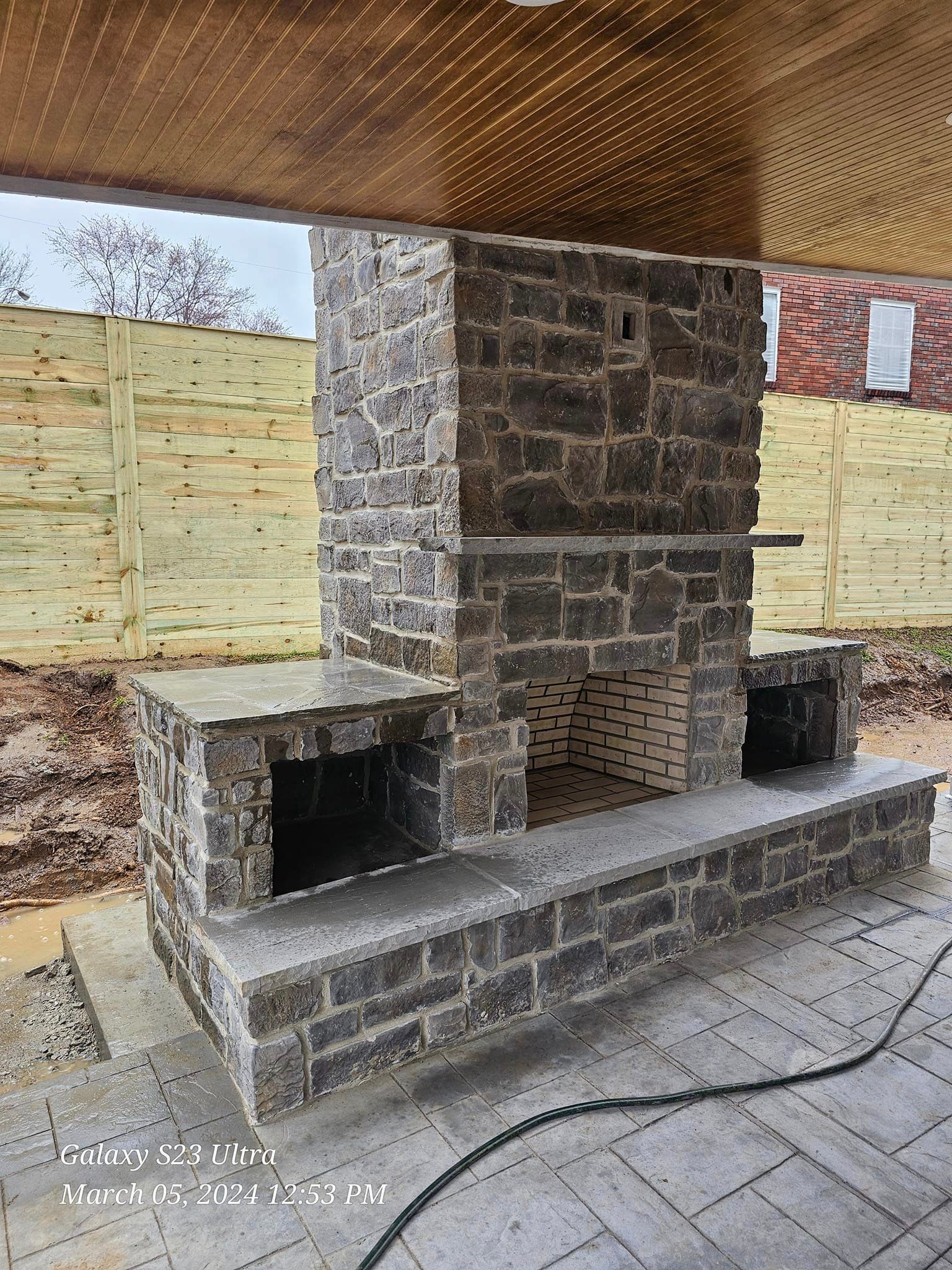 Stone outdoor fireplace with a brick facade and gray accents, under a wooden ceiling.