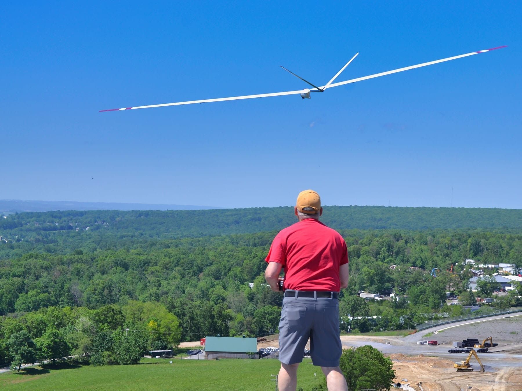 Man operating a long-winged glider with a remote control, blue sky above green trees.