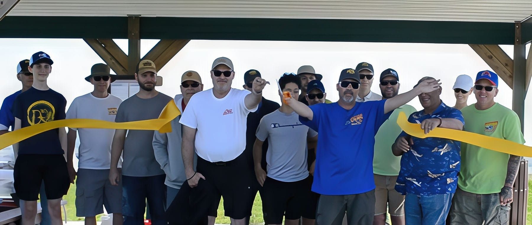 A group of people cutting a yellow ribbon under a wooden shelter, celebrating an opening.