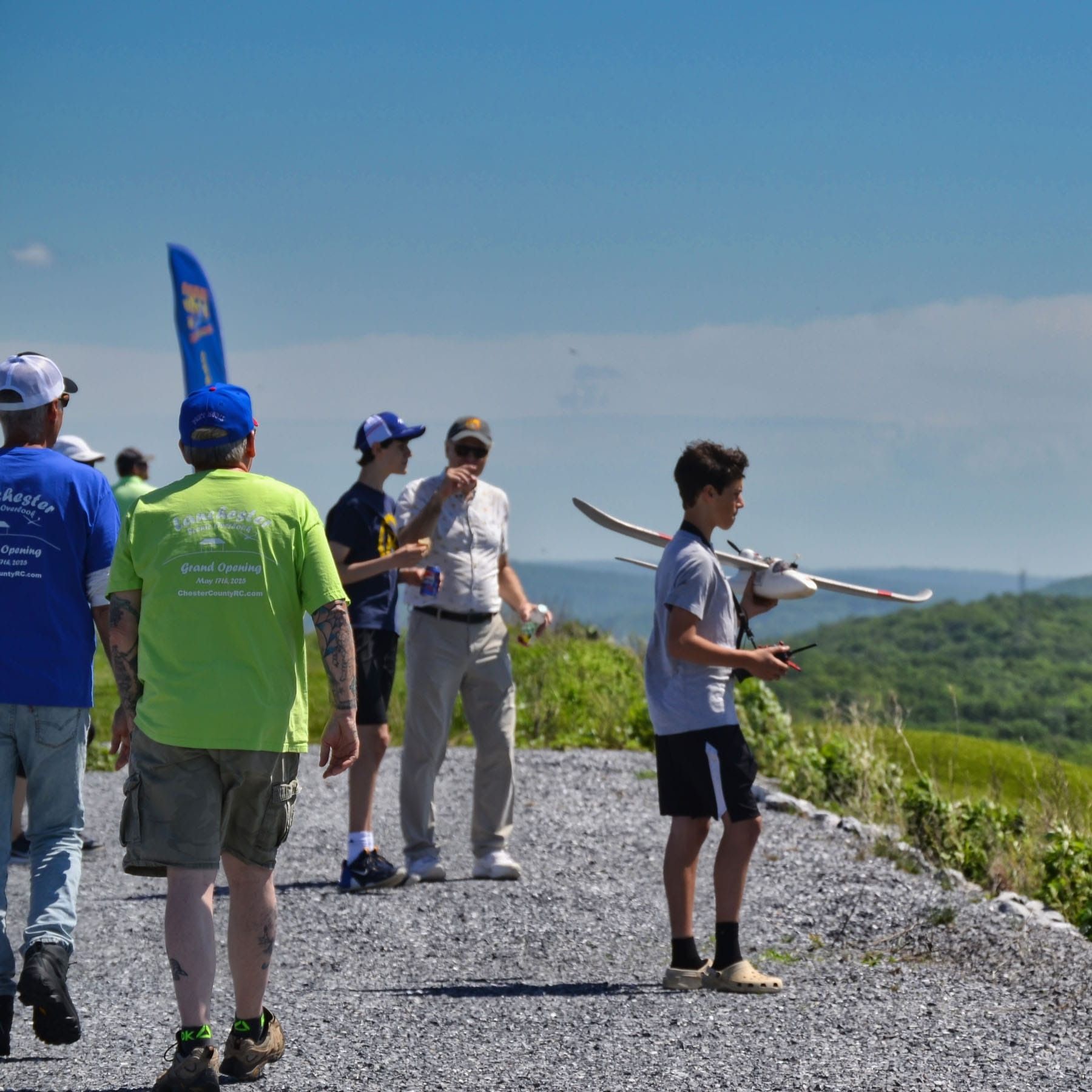 Group watches boy fly a model airplane on a gravel path overlooking a green valley under a blue sky.