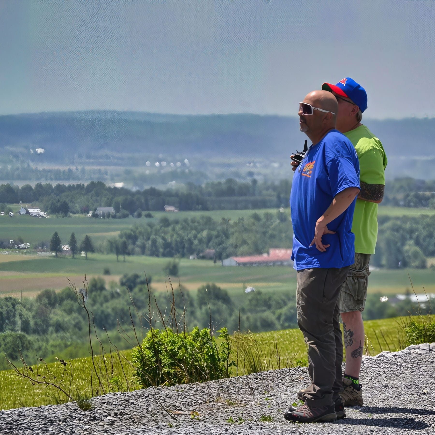 Two men overlooking a valley from a hilltop. One in blue shirt, other in green, both wearing hats. Scenic view.