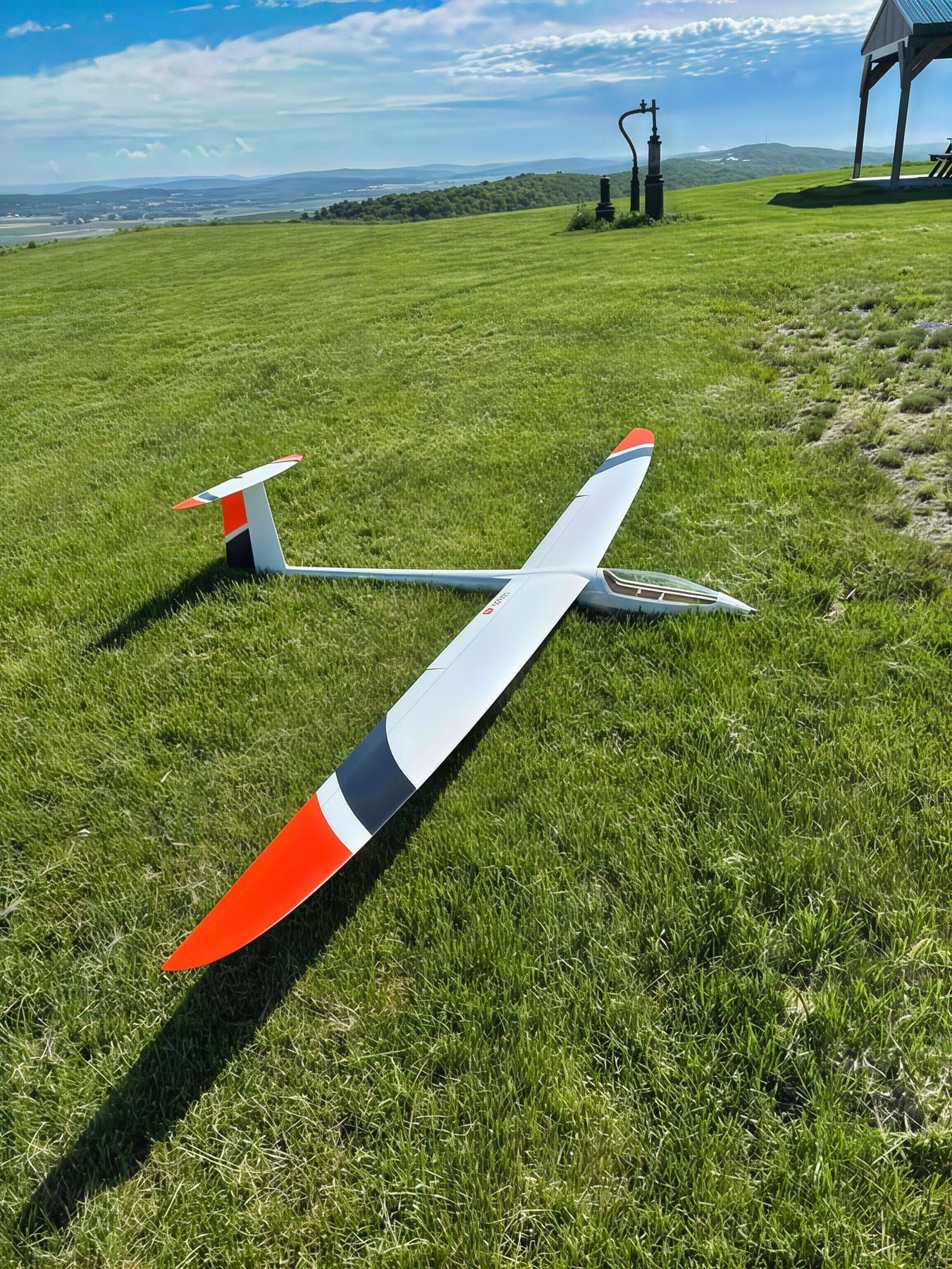 White and orange glider on green grass, with wings spread. Blue sky and hilltop background.