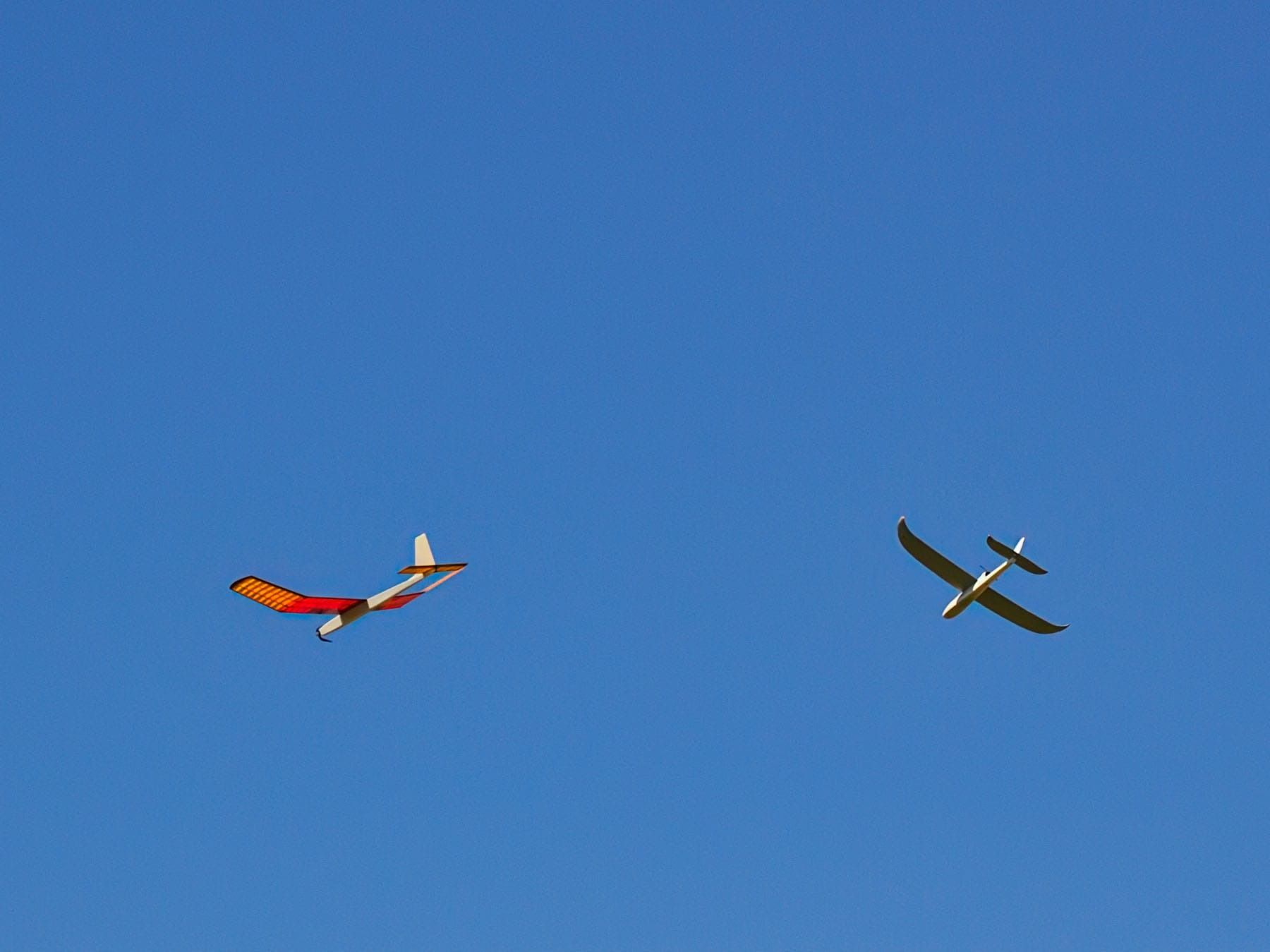 Two model airplanes, one red and yellow, the other white, flying in a clear blue sky.