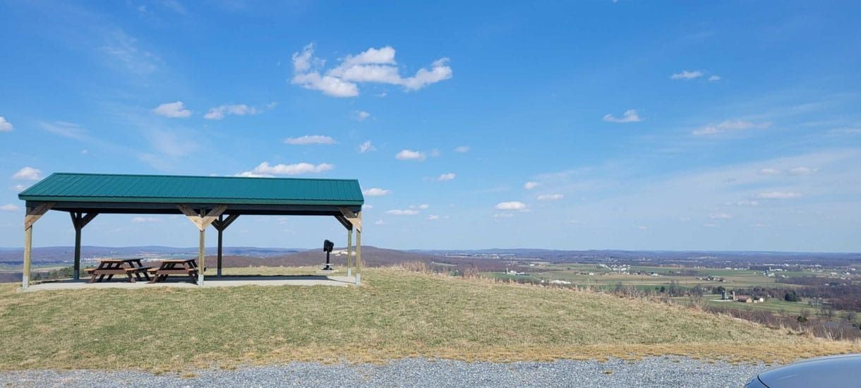 A green-roofed shelter on a grassy hilltop overlooks a distant landscape under a blue sky with clouds.
