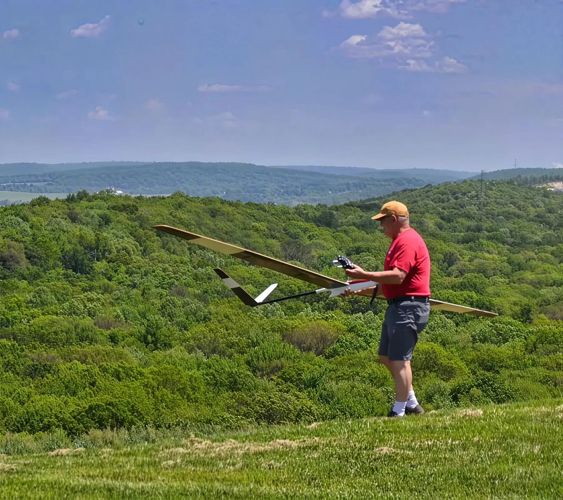 Man in red shirt launching a glider from a grassy hilltop overlooking a forested valley.