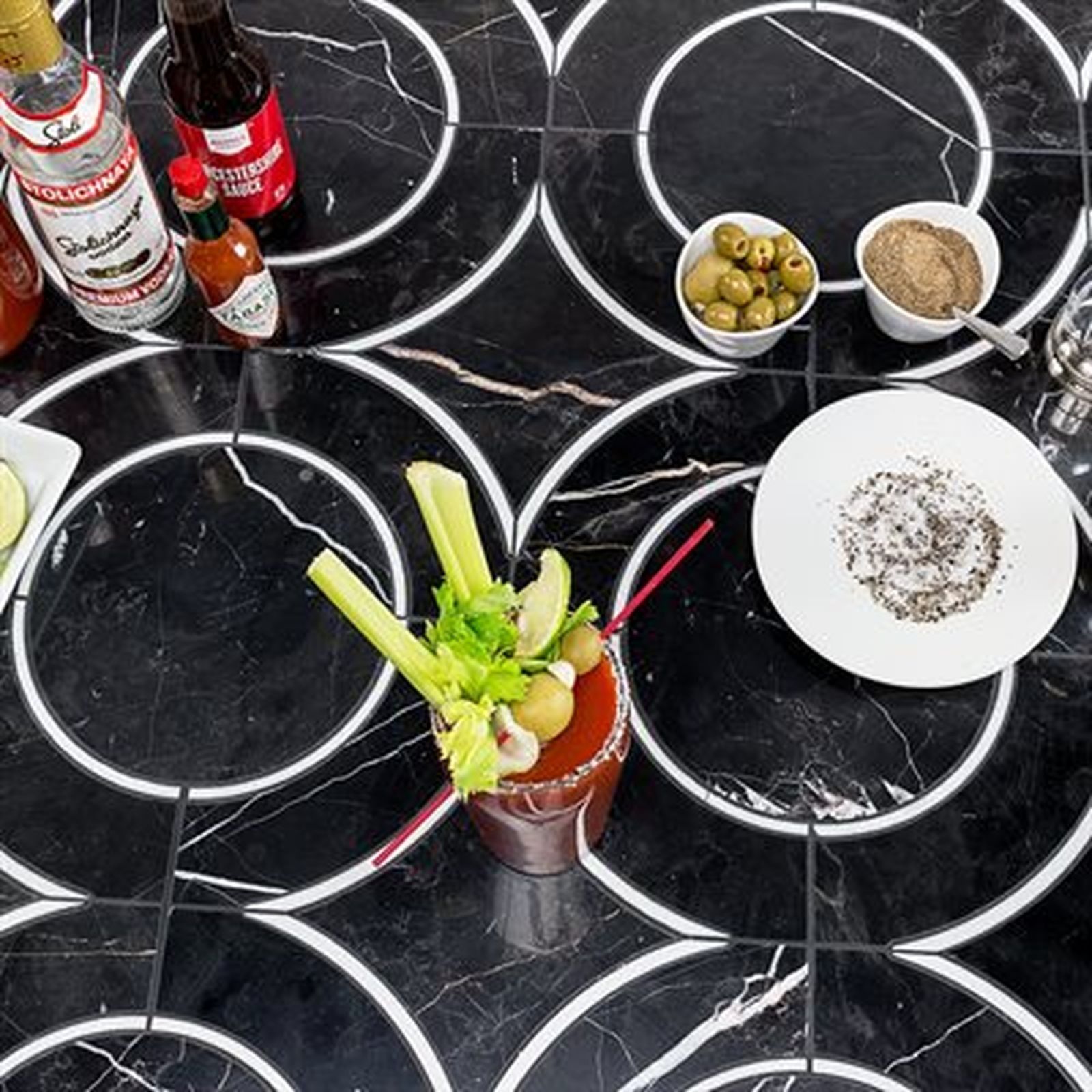A Bloody Mary cocktail with garnishes, ingredients on a black tile surface.