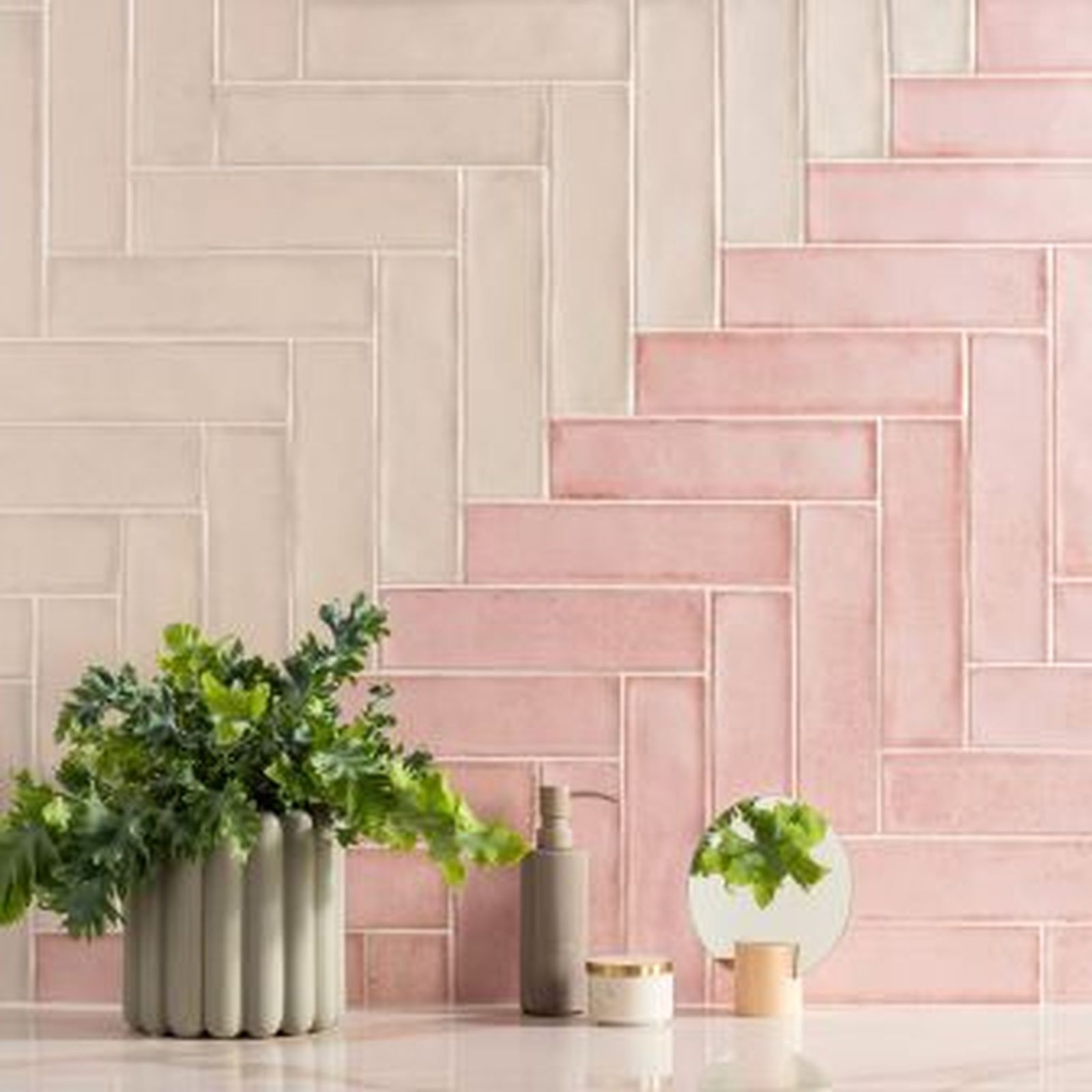 Herringbone tile wall in light and medium pink, with toiletries and a potted plant in front.